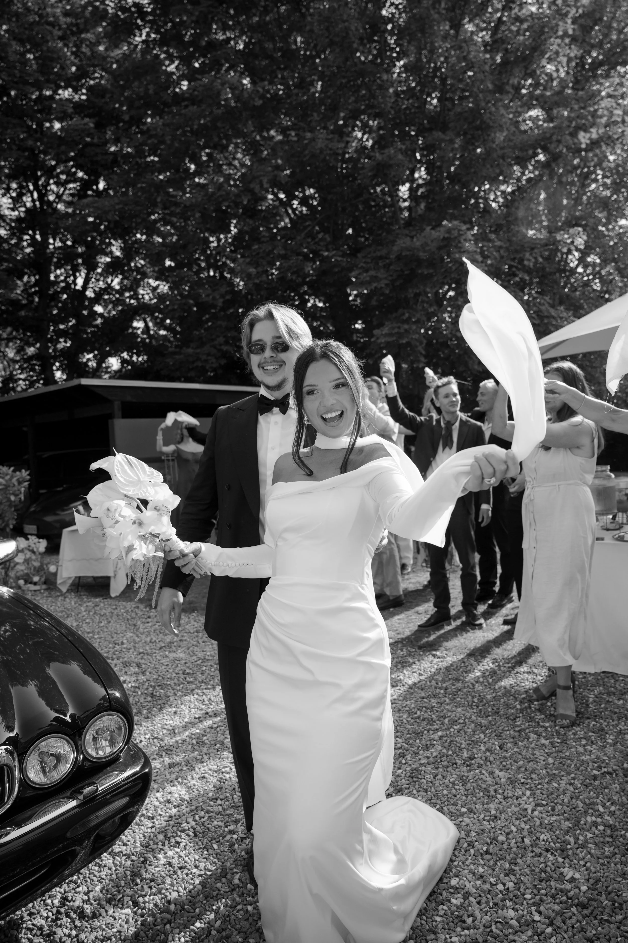 Black and white photo of a bride and groom celebrating outdoors with friends, wedding reception in background, bride holding a bouquet and waving a cloth.