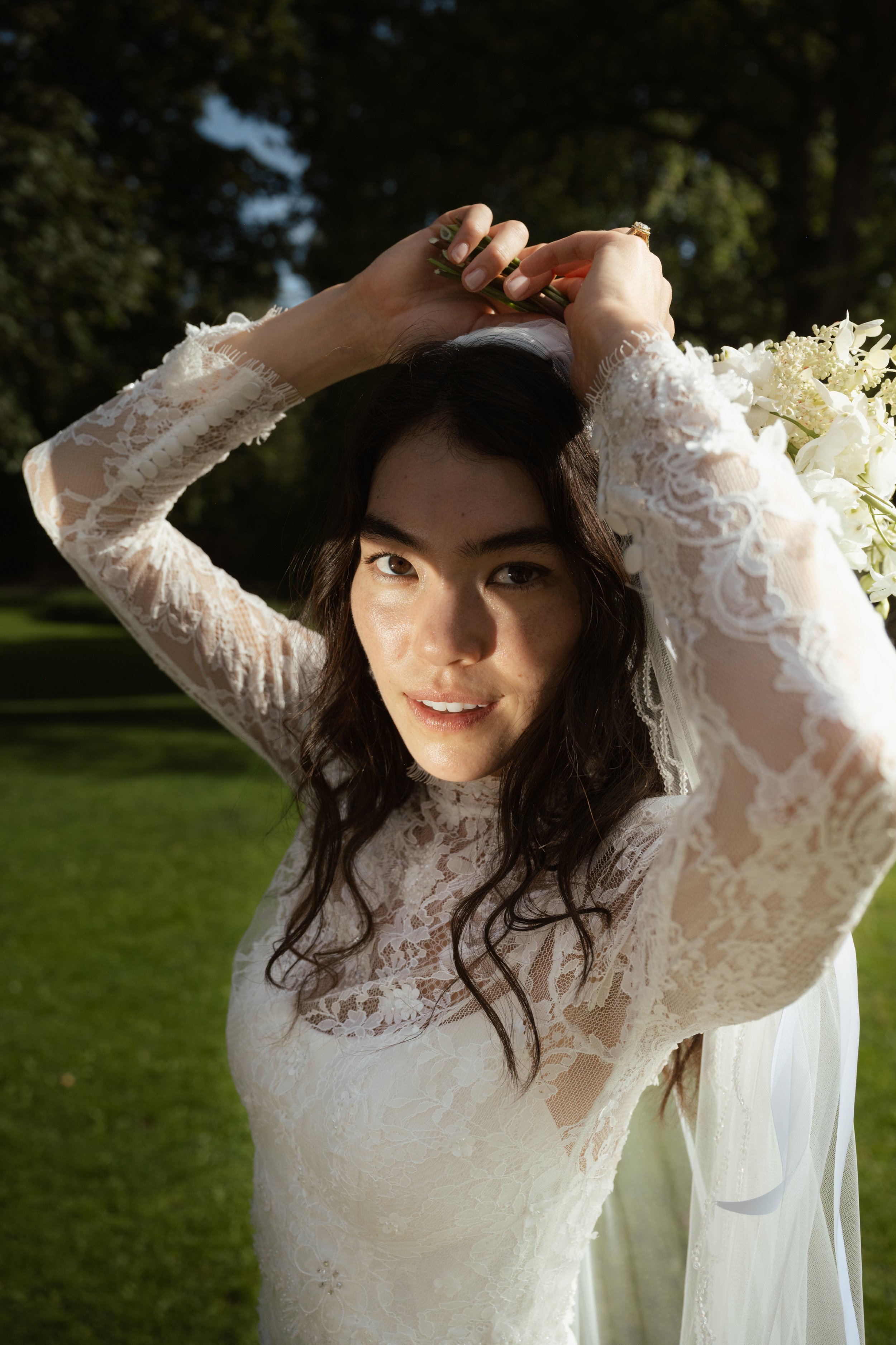 A woman in a white lace dress with long sleeves is outdoors on a sunny day, lifting a small bouquet of flowers above her head, with green trees and grass in the background.