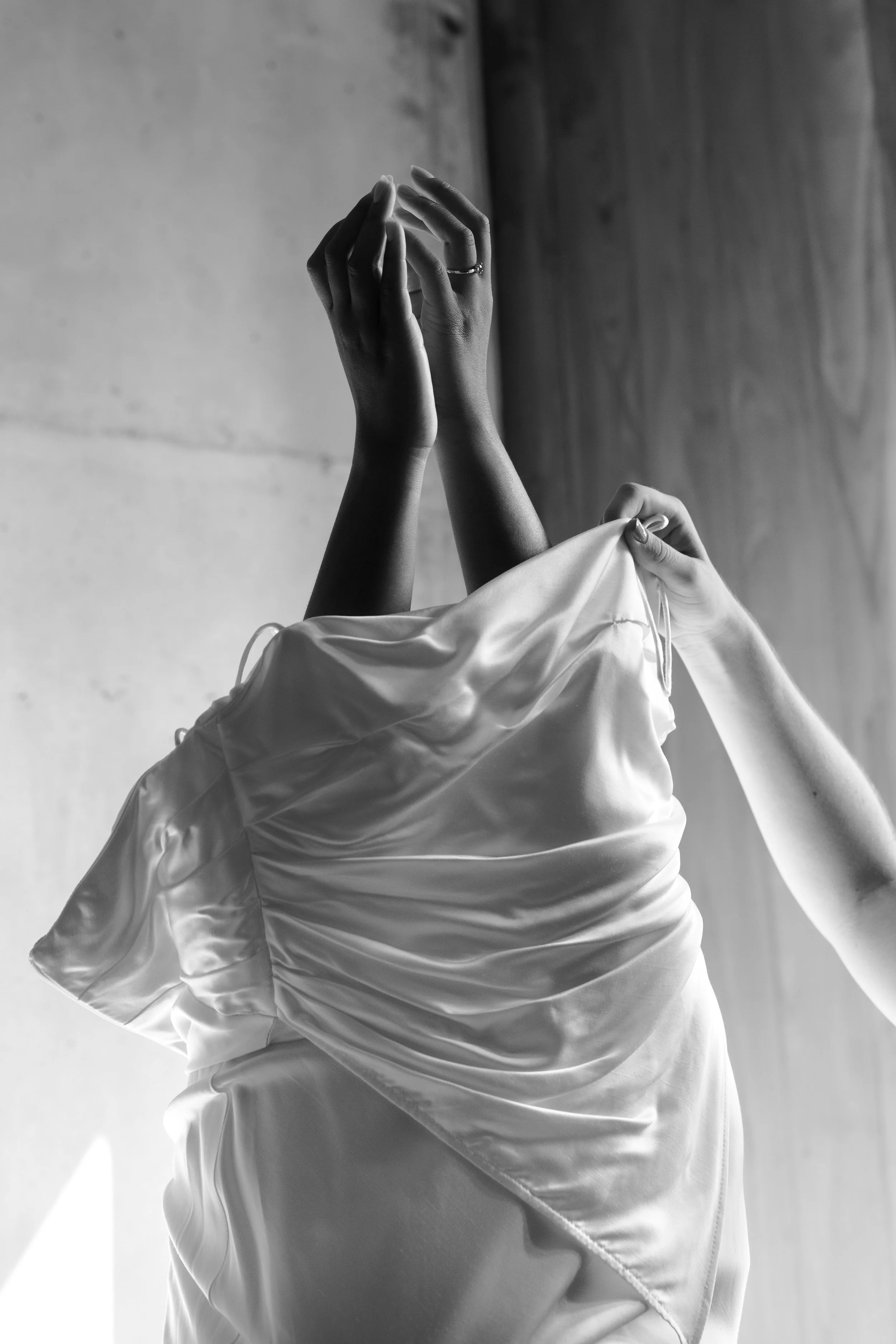 Black and white photograph of a person with hands raised, wearing a satin dress, with a textured background.