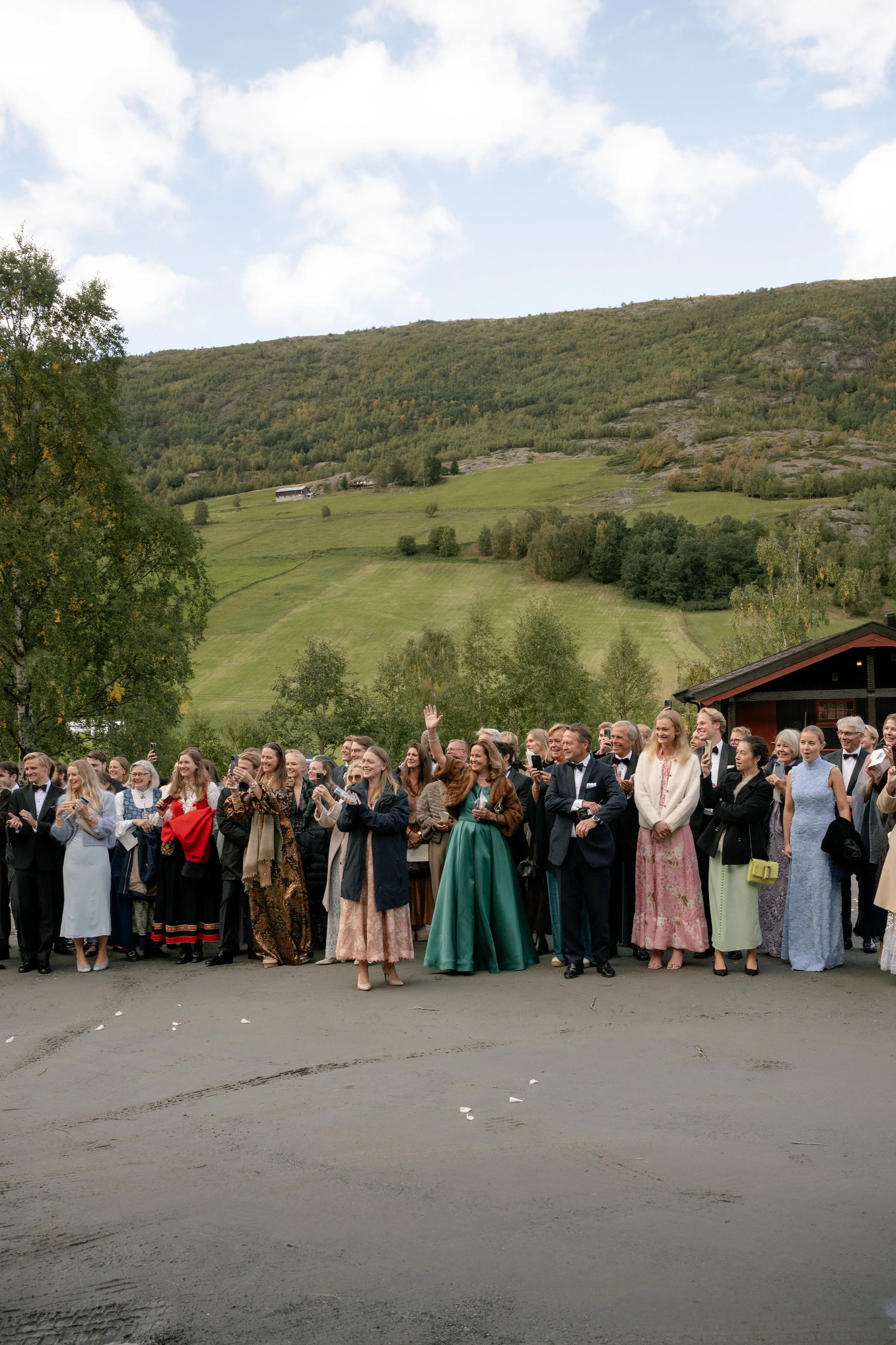 Group of well-dressed people, including men in tuxedos and women in dresses, gathered outdoors with scenic green hills and a cloudy sky in the background.