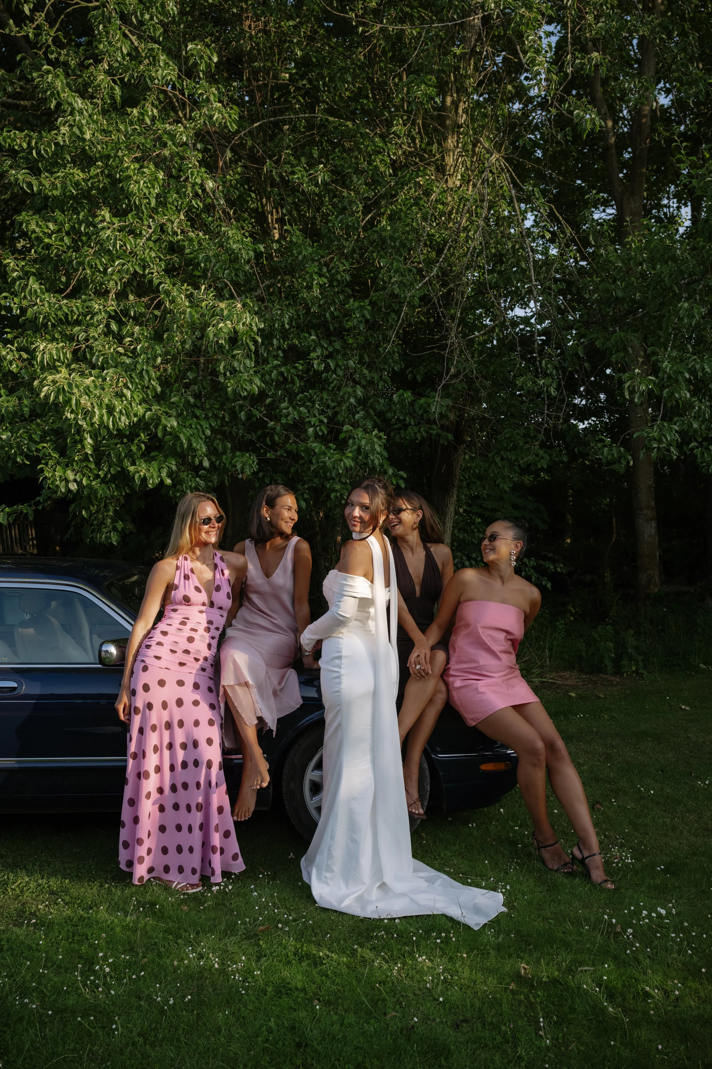 A group of five women in colorful dresses posing next to a black car outdoors with green trees in the background.