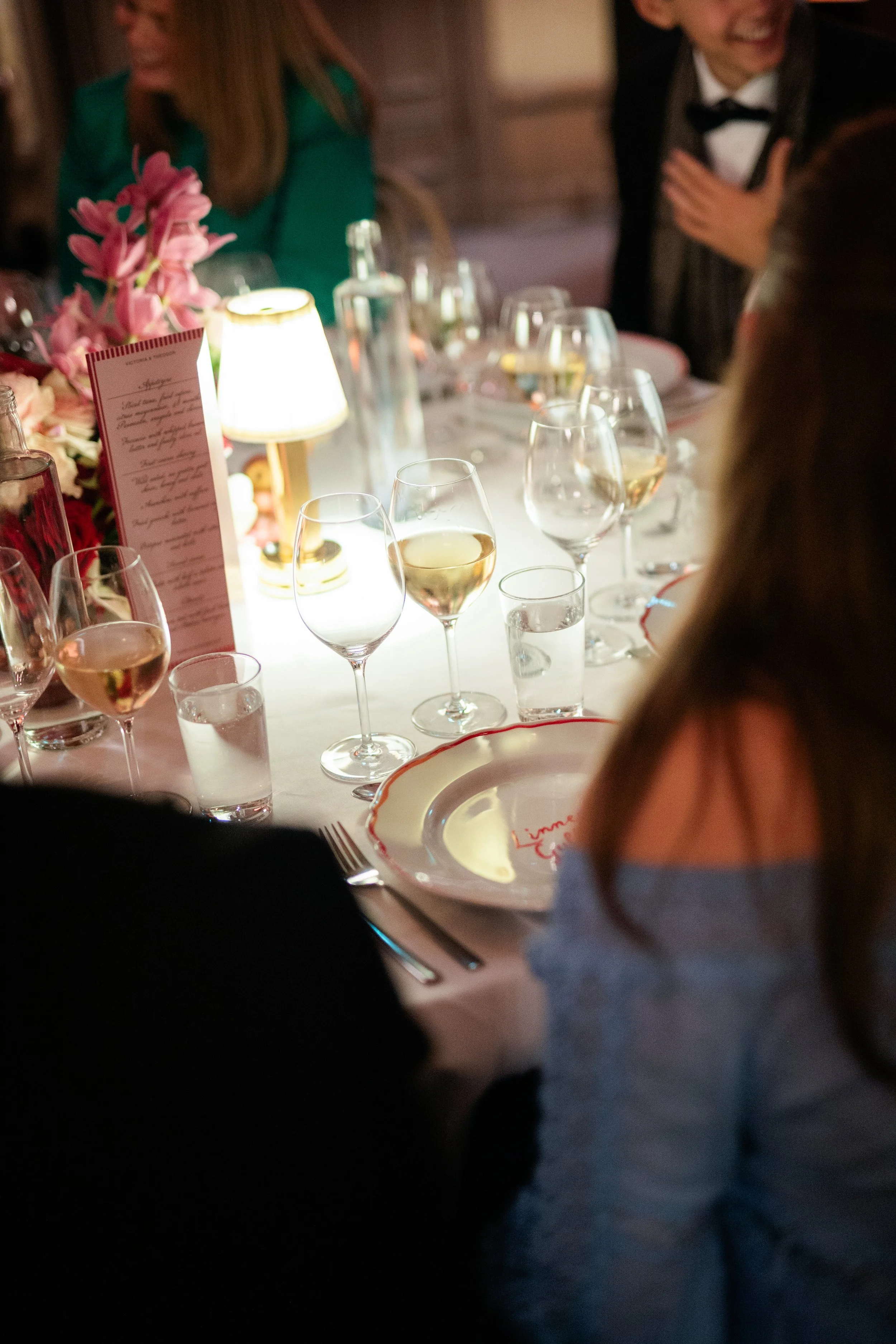 People gathered around a dining table with glasses of white wine, a pink flower centerpiece, a small lamp, and a menu, at a formal event or celebration.