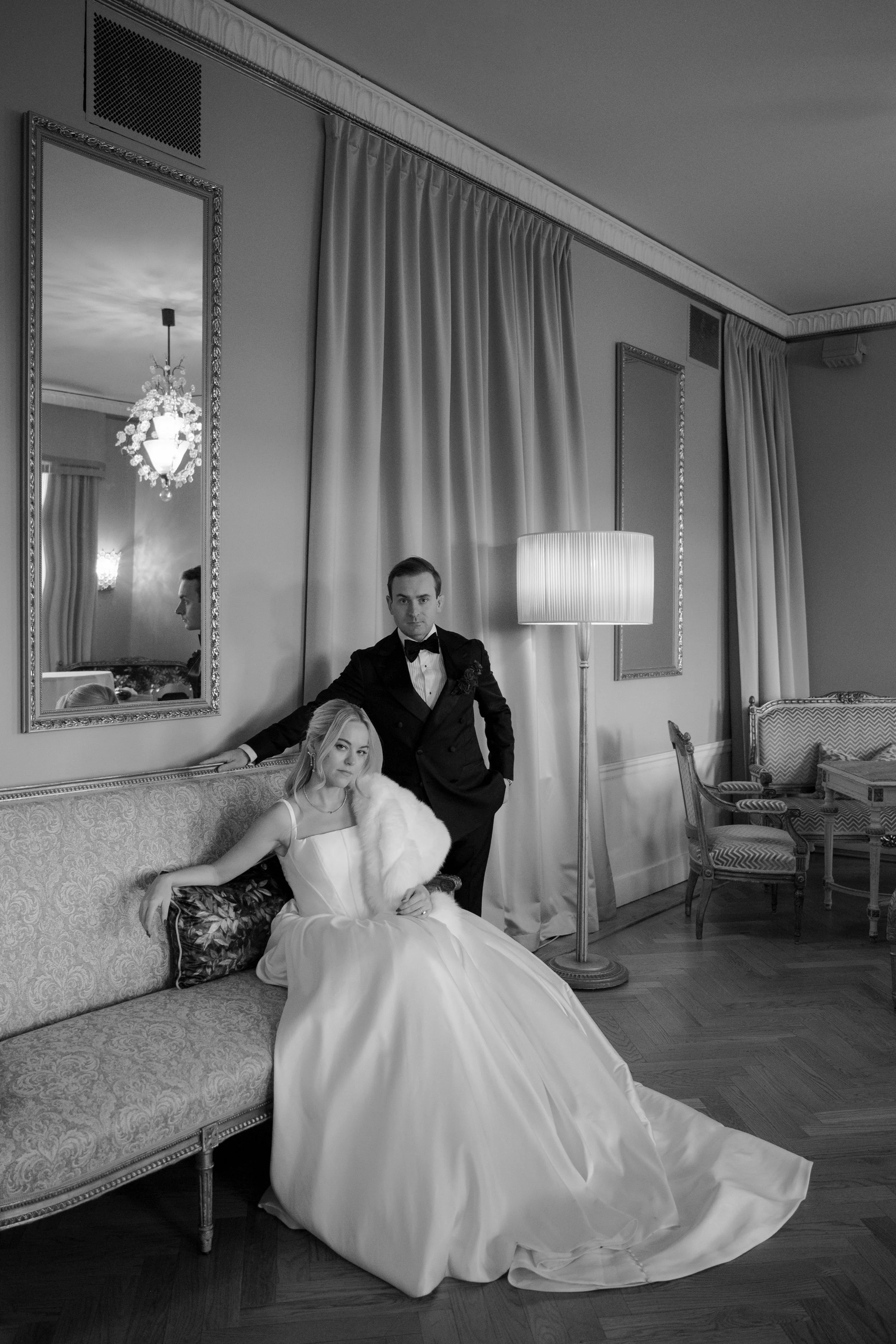 A black and white photo of a bride in a wedding gown sitting on a vintage sofa, with a groom in a tuxedo standing beside her in an elegant room with curtains, mirrors, a lamp, and antique furniture.