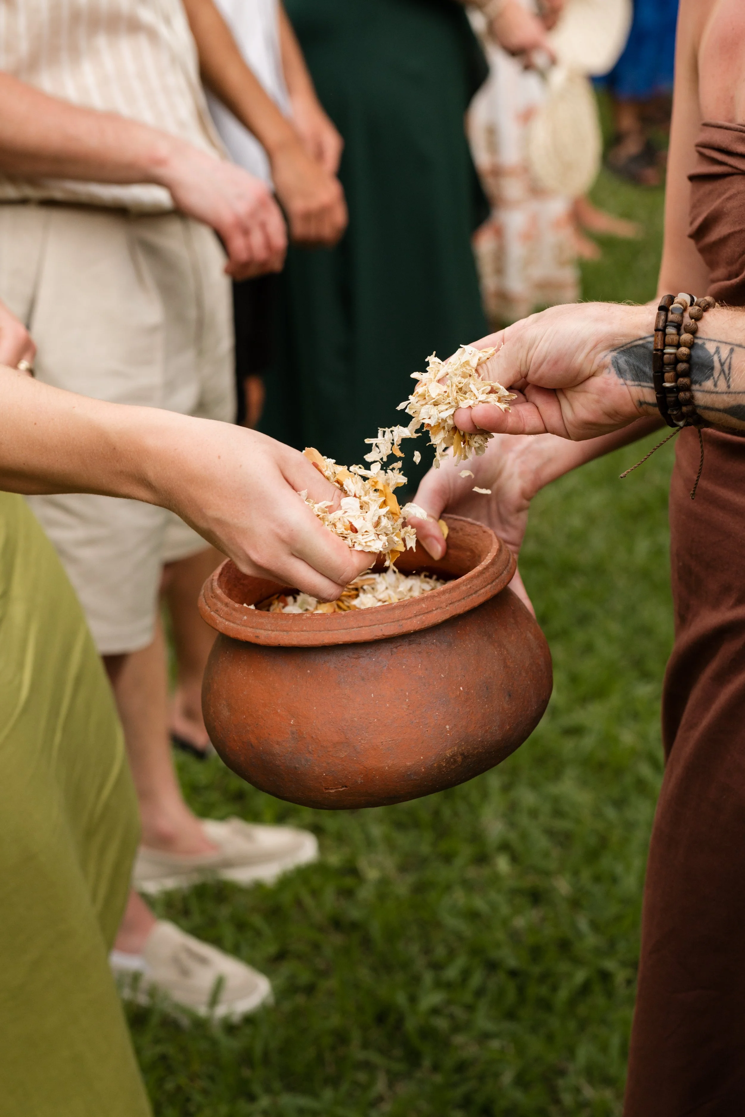 People participating in a traditional ritual, placing or exchanging small objects or offerings into a clay pot, outdoors on grass.