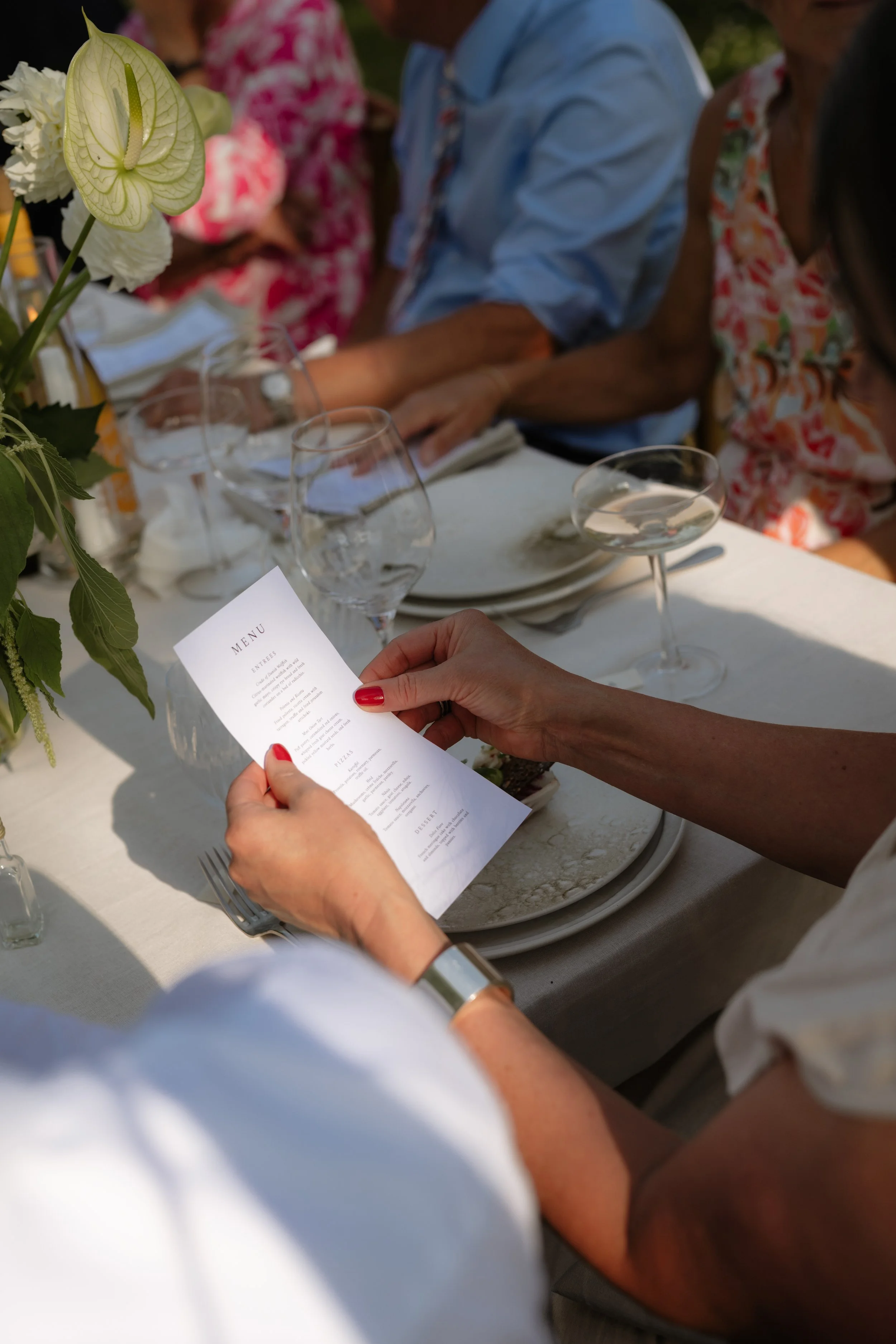 A person holding a menu at a table set for a meal, with glassware and a flower arrangement on the table, while other guests sit nearby.