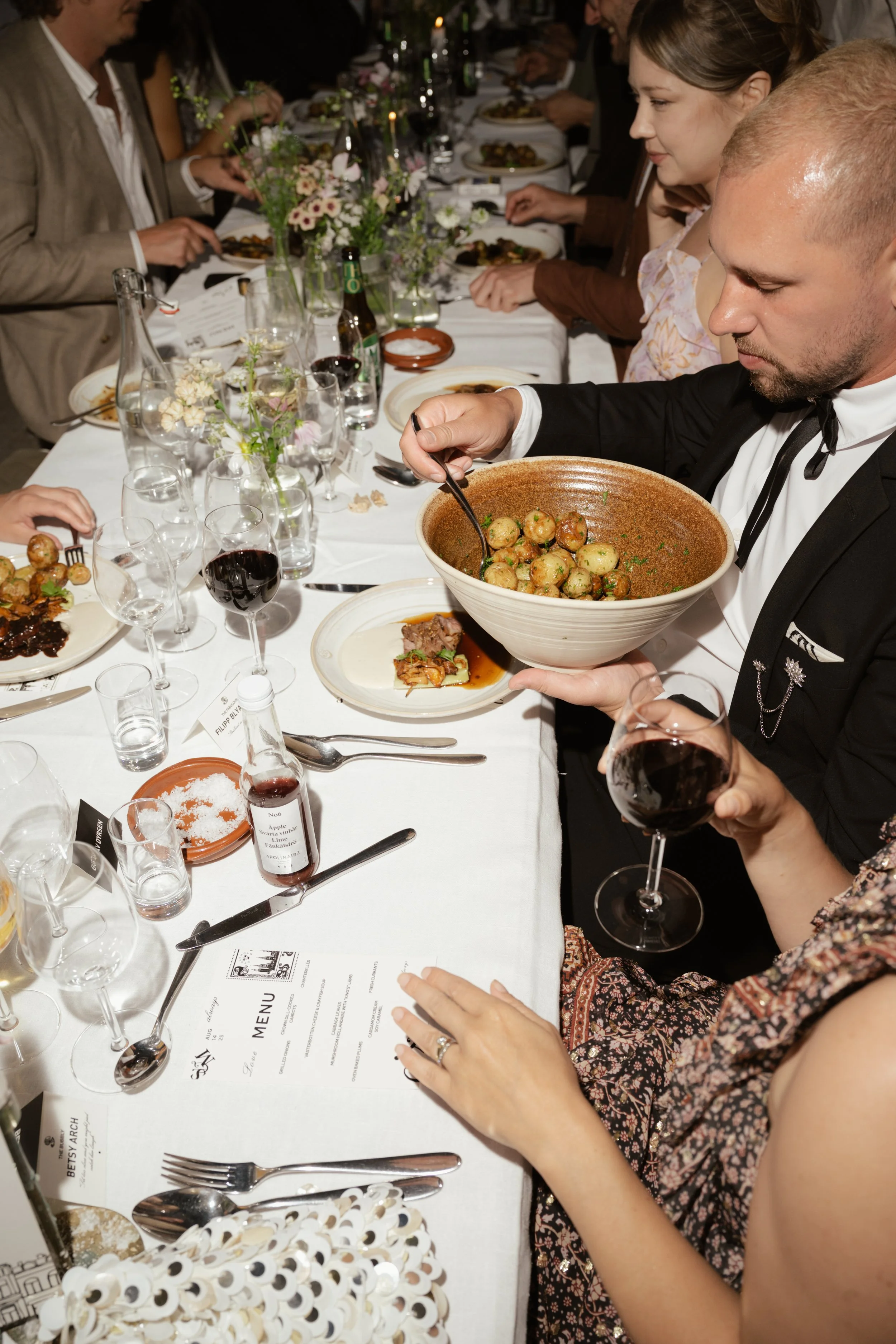 People dining at a formal dinner party, with wine glasses, plates, flowers, and a man serving food from a large bowl.