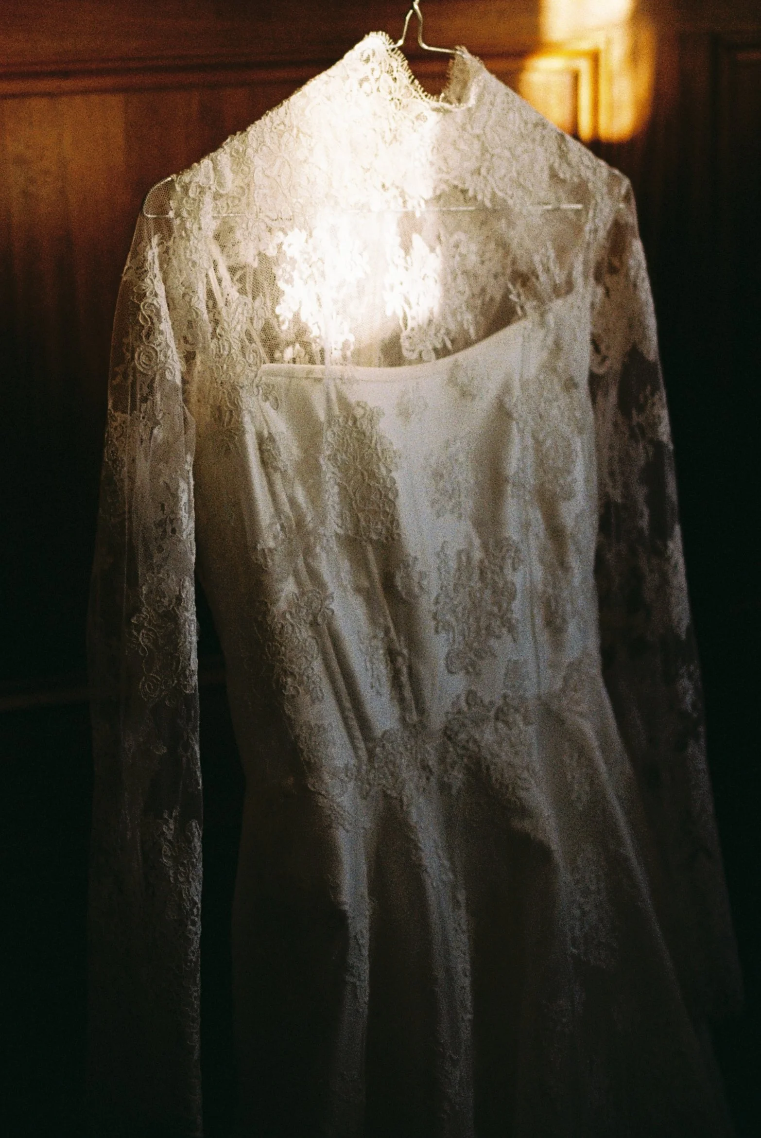 A white lace wedding dress hanging on a wooden hanger in a dimly lit room.