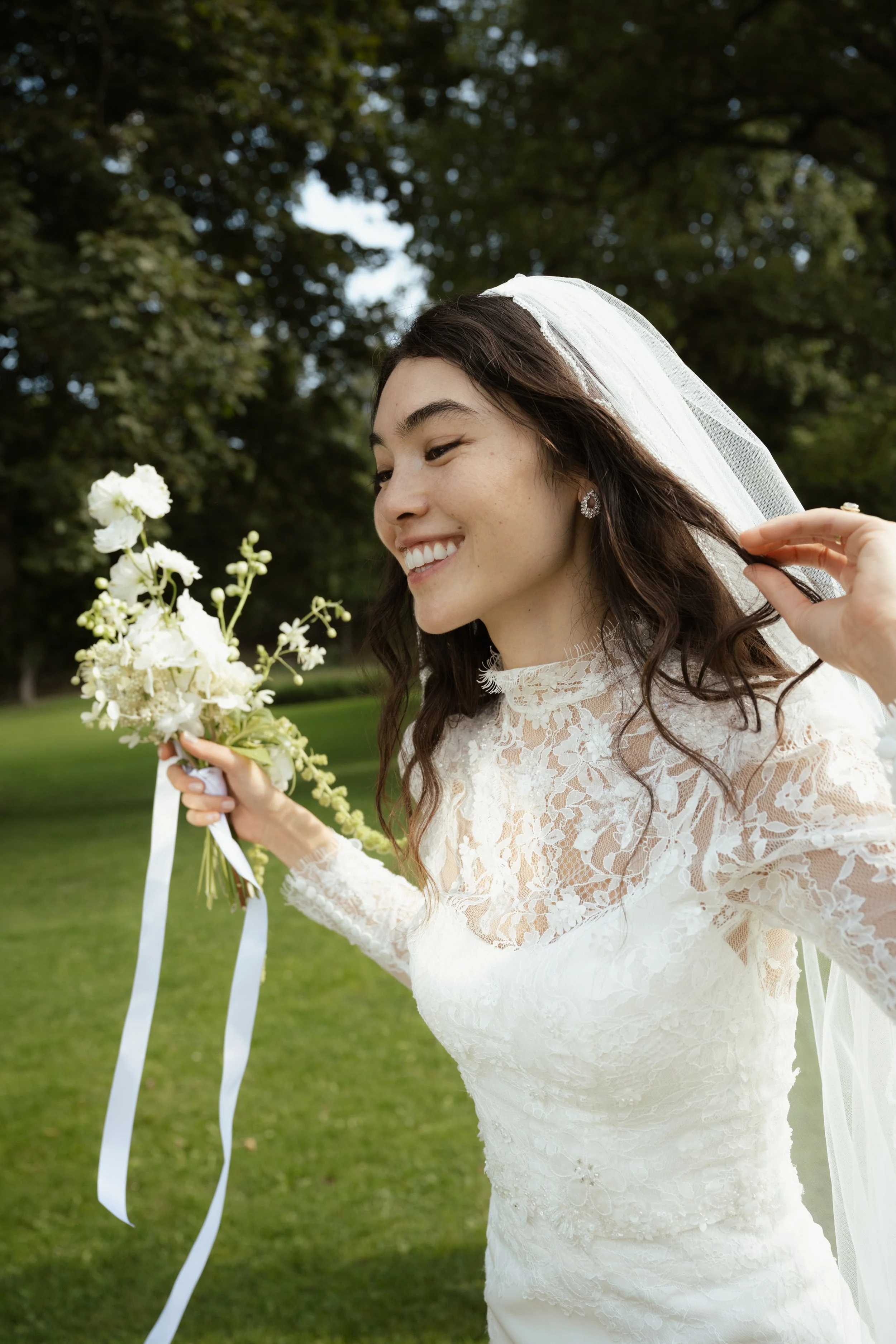 A bride in a lace wedding dress holding a bouquet of white flowers outdoors with trees in the background.