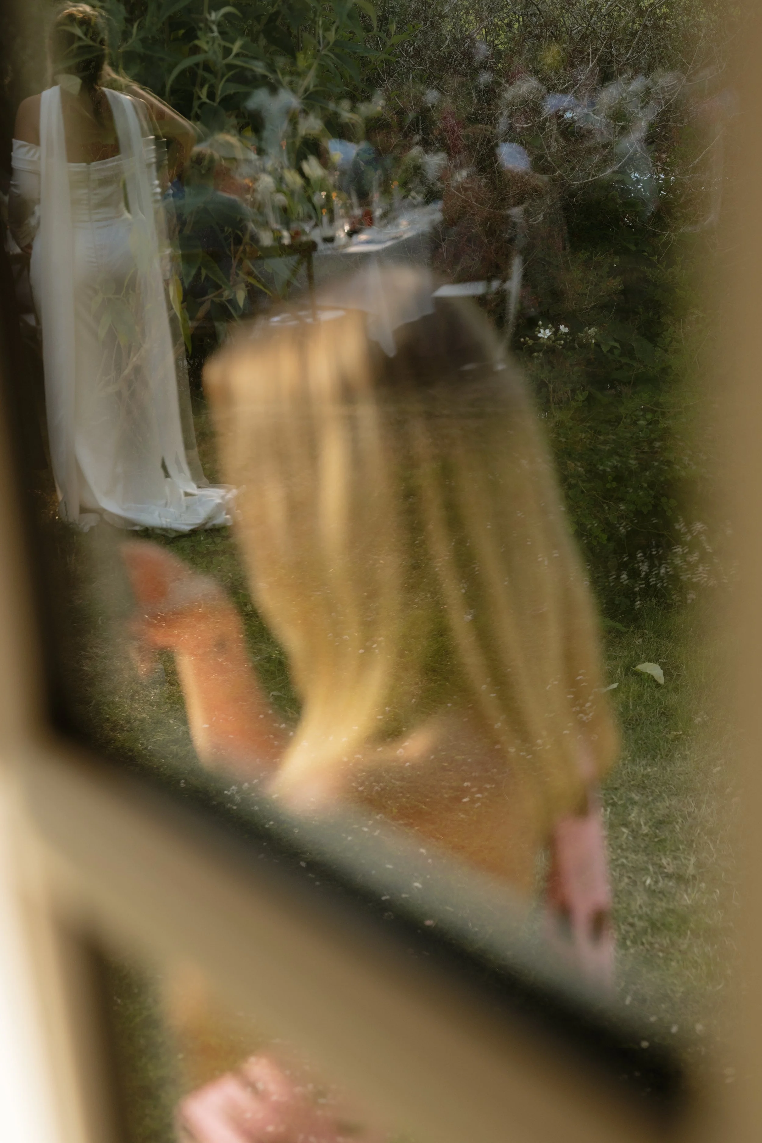 A woman in a white dress seen through a window with reflections, during an outdoor gathering or event with tables and seated guests.