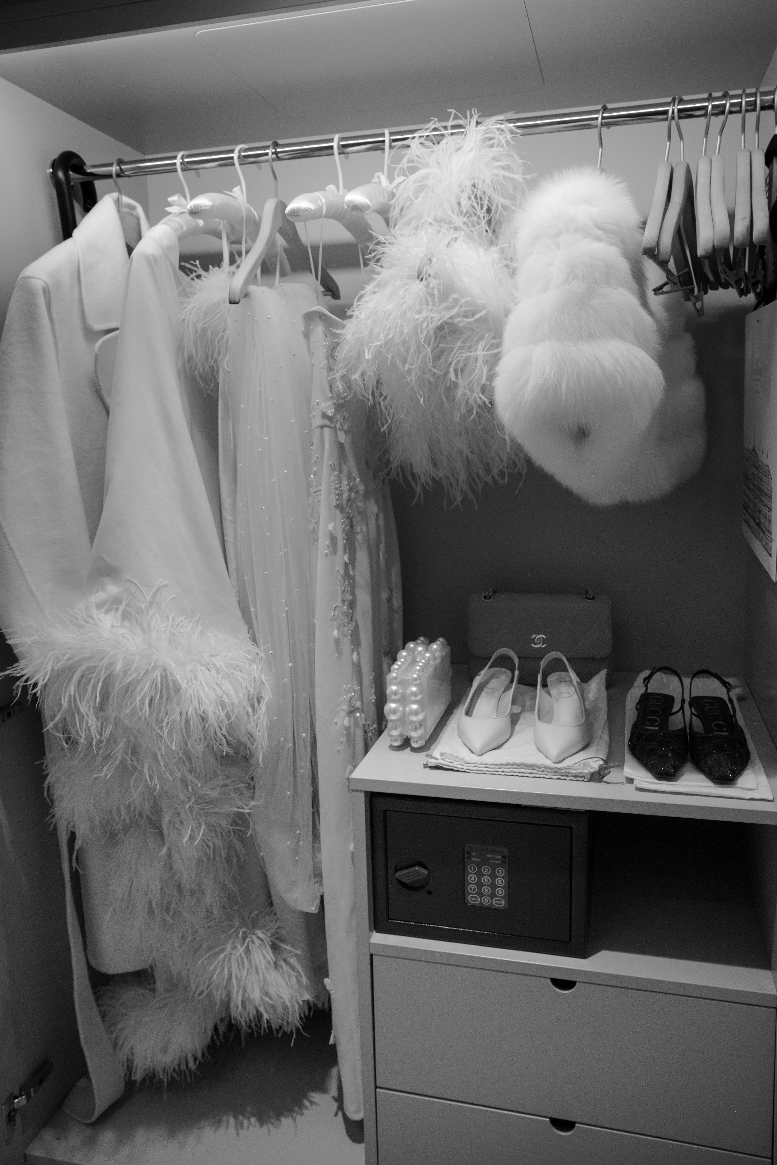 Closet with white clothing and feathered accessories, and a small table with shoes, a pearl clutch, and a safe.