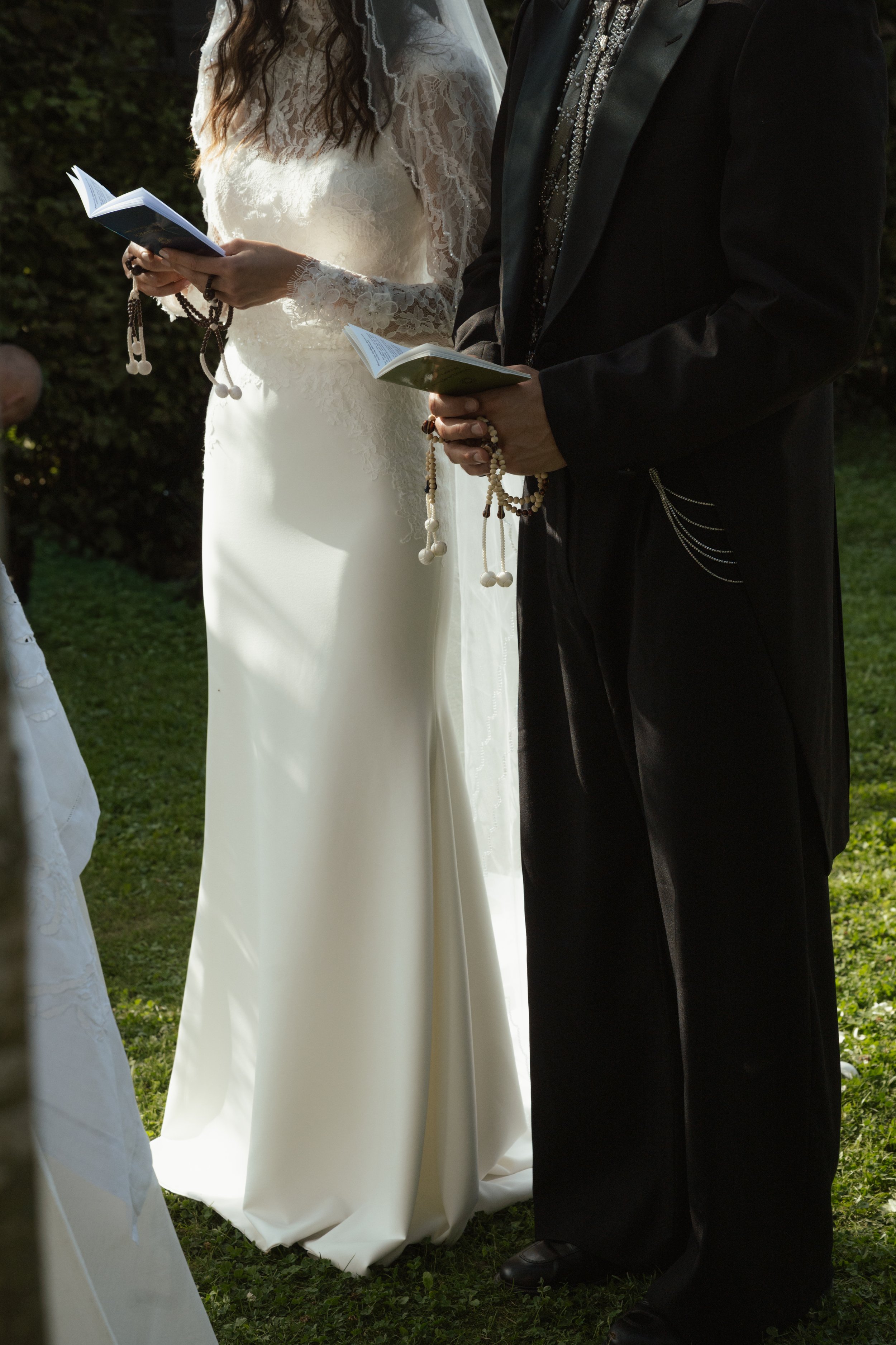 A bride in a white lace wedding dress and a groom in a black tuxedo standing outdoors during a wedding ceremony, each holding a small book or booklet, likely for their vows.