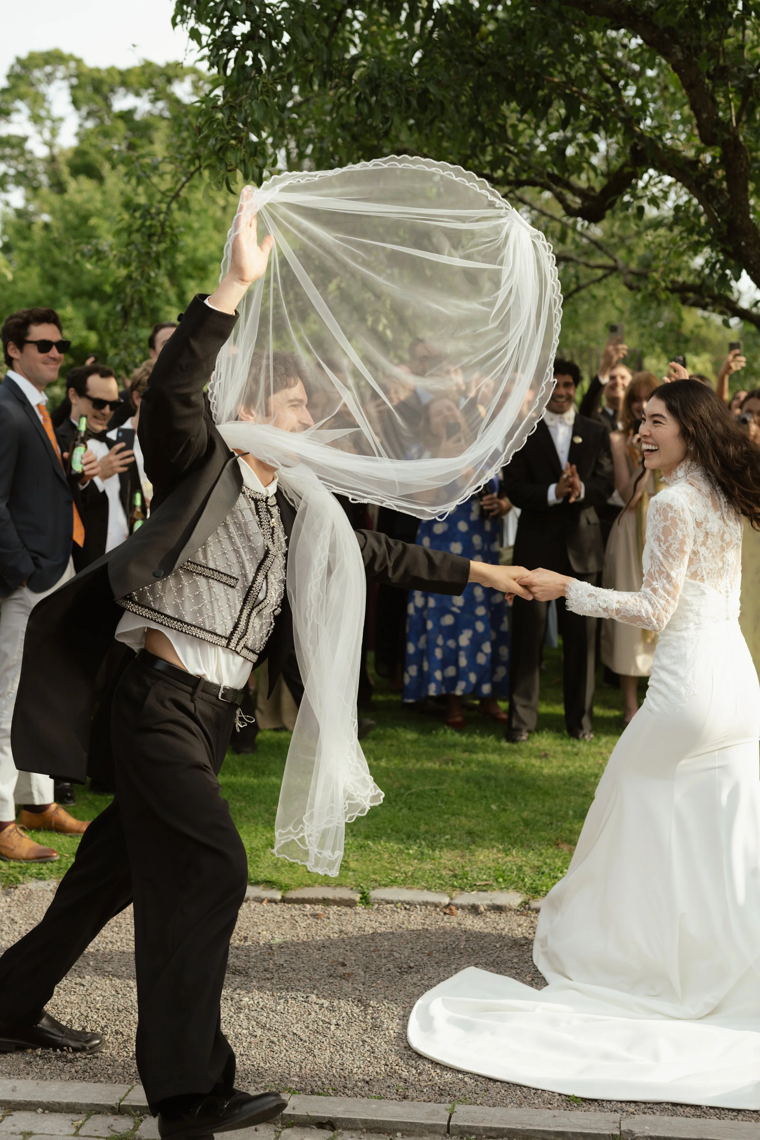 A wedding ceremony outdoors with a groom in a tuxedo lifting a sheer veil over the bride in a lace wedding dress while they hold hands, surrounded by guests in formal attire.