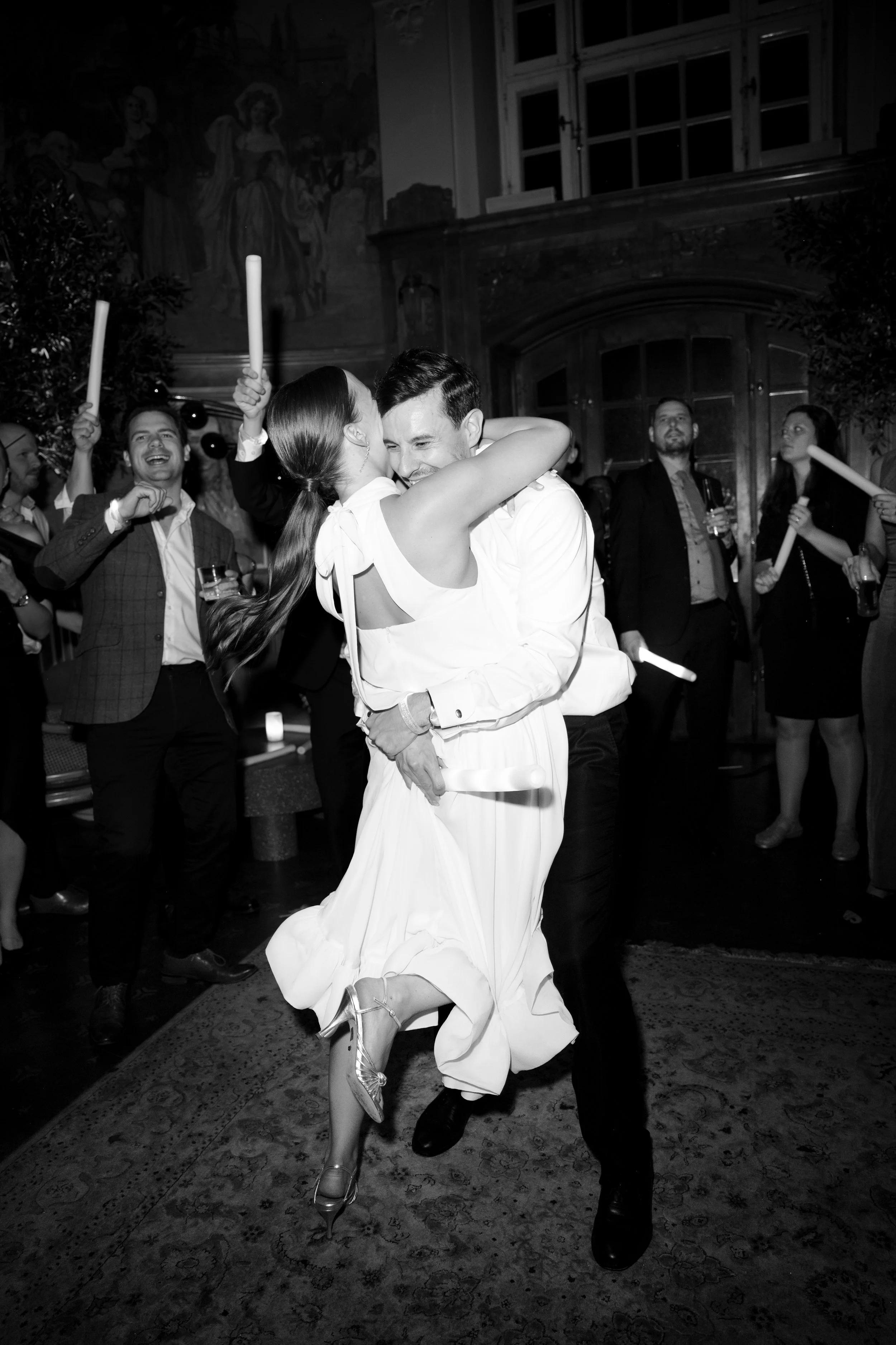 A couple dancing closely at a wedding reception in a decorated indoor venue, with guests holding candles or drinks in the background.