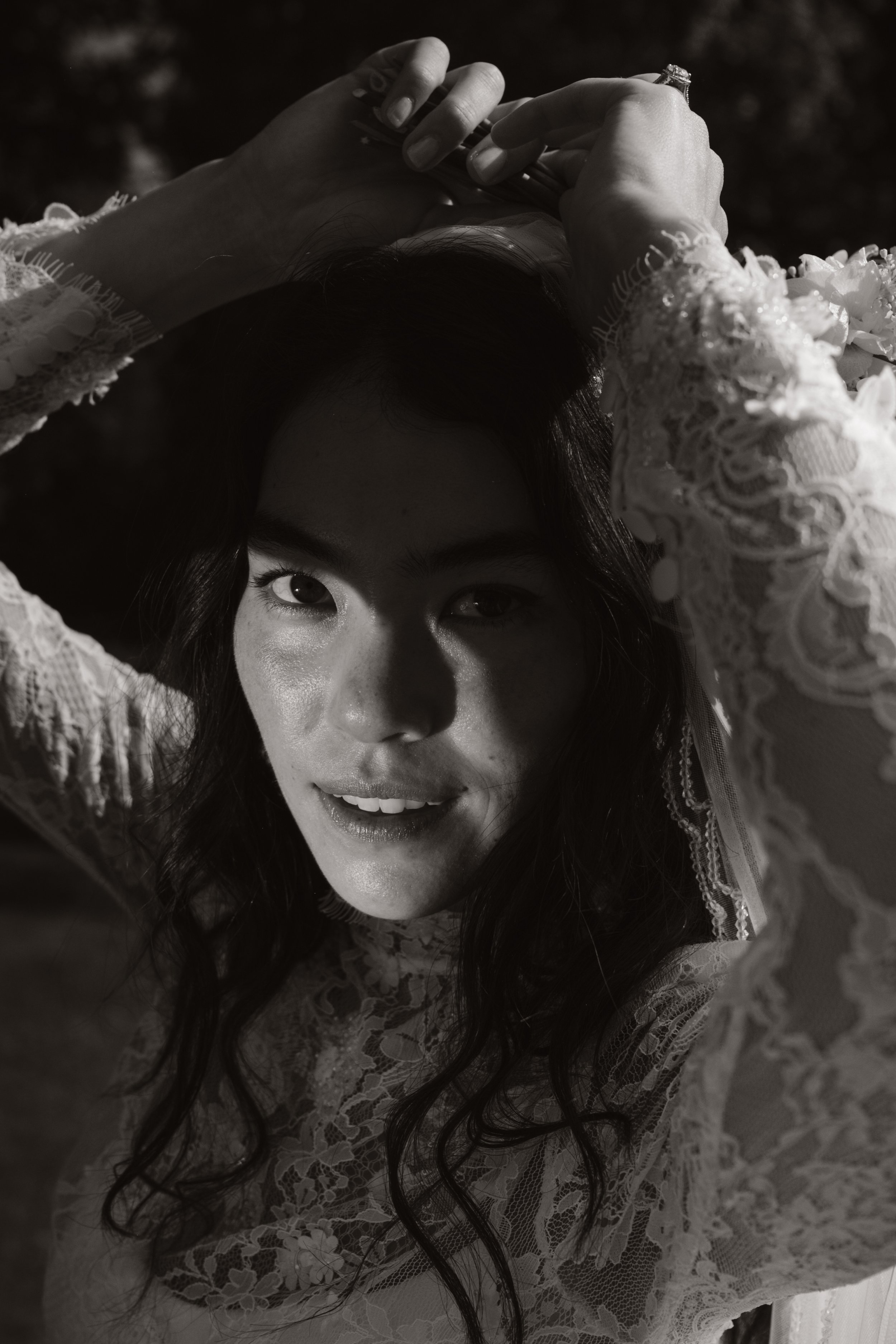 Black and white photo of a woman with dark hair wearing a lace dress, posing outdoors with her arms raised above her head, looking directly at the camera.