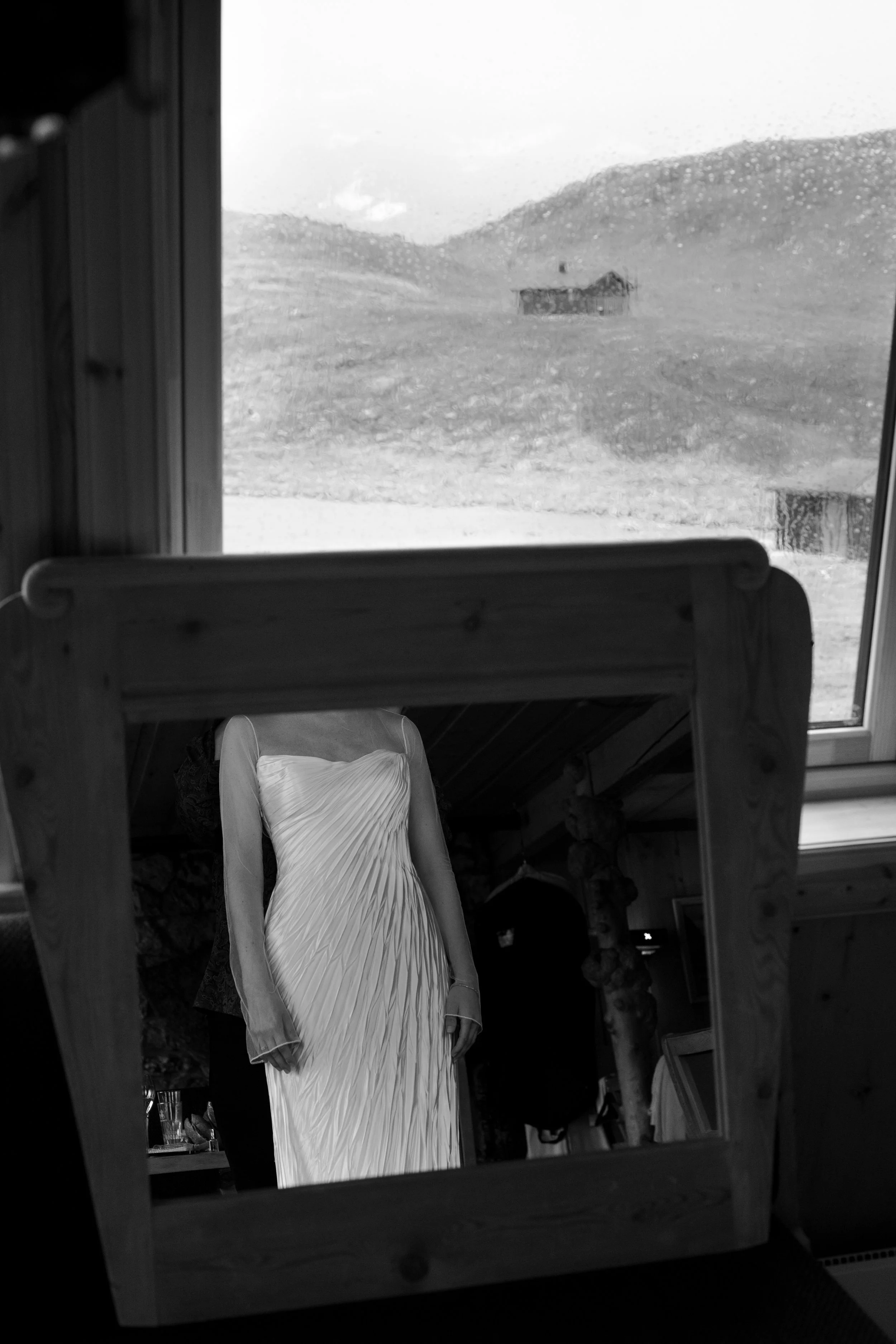 A woman in a strapless, pleated white dress looking at her reflection in a wooden-framed mirror, with a landscape view of hills and a house seen through a rain-covered window in the background.