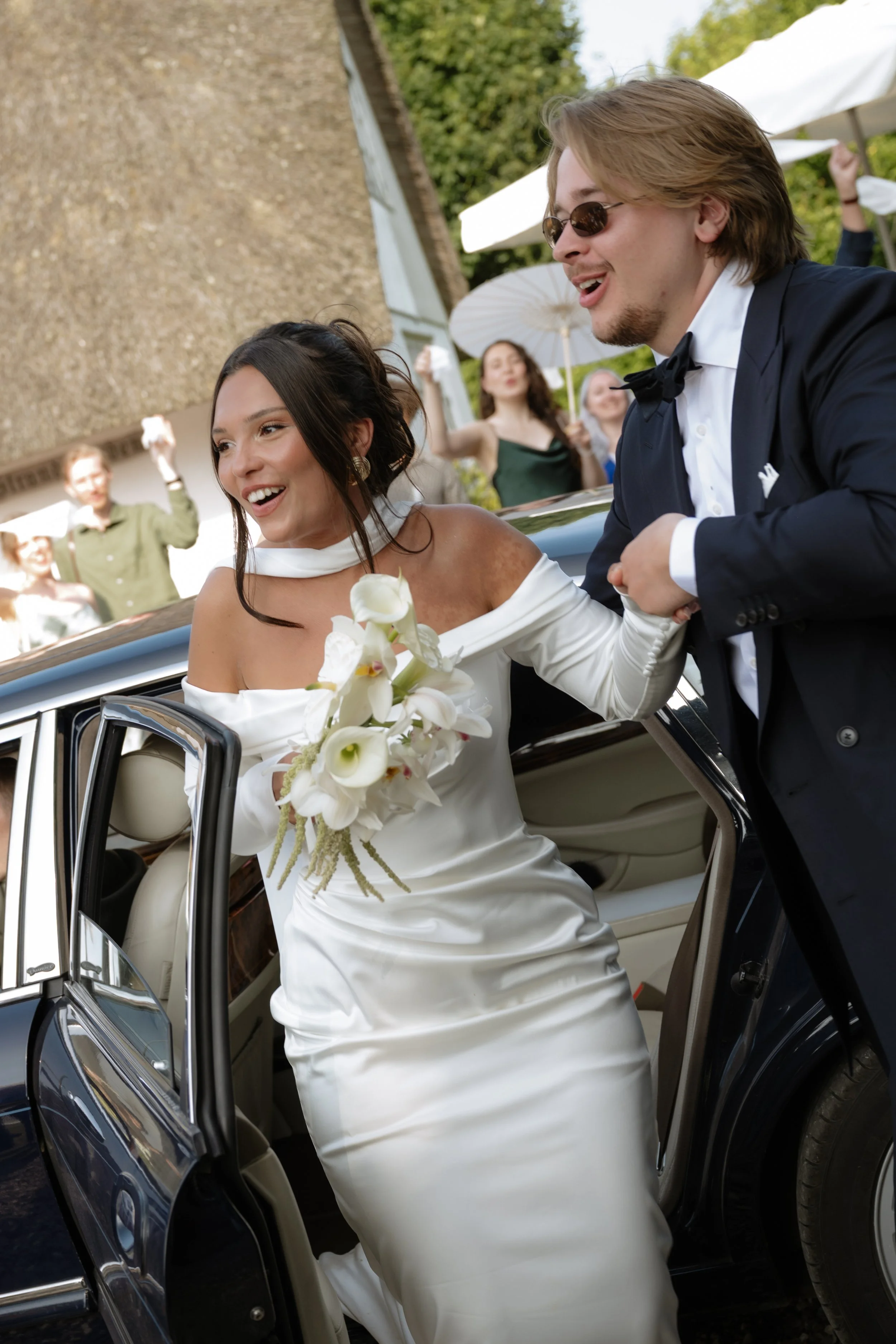 Bride in white wedding gown holding a bouquet of white flowers getting into a car at her wedding, groom in a black tuxedo helping her, guests with umbrellas in the background.