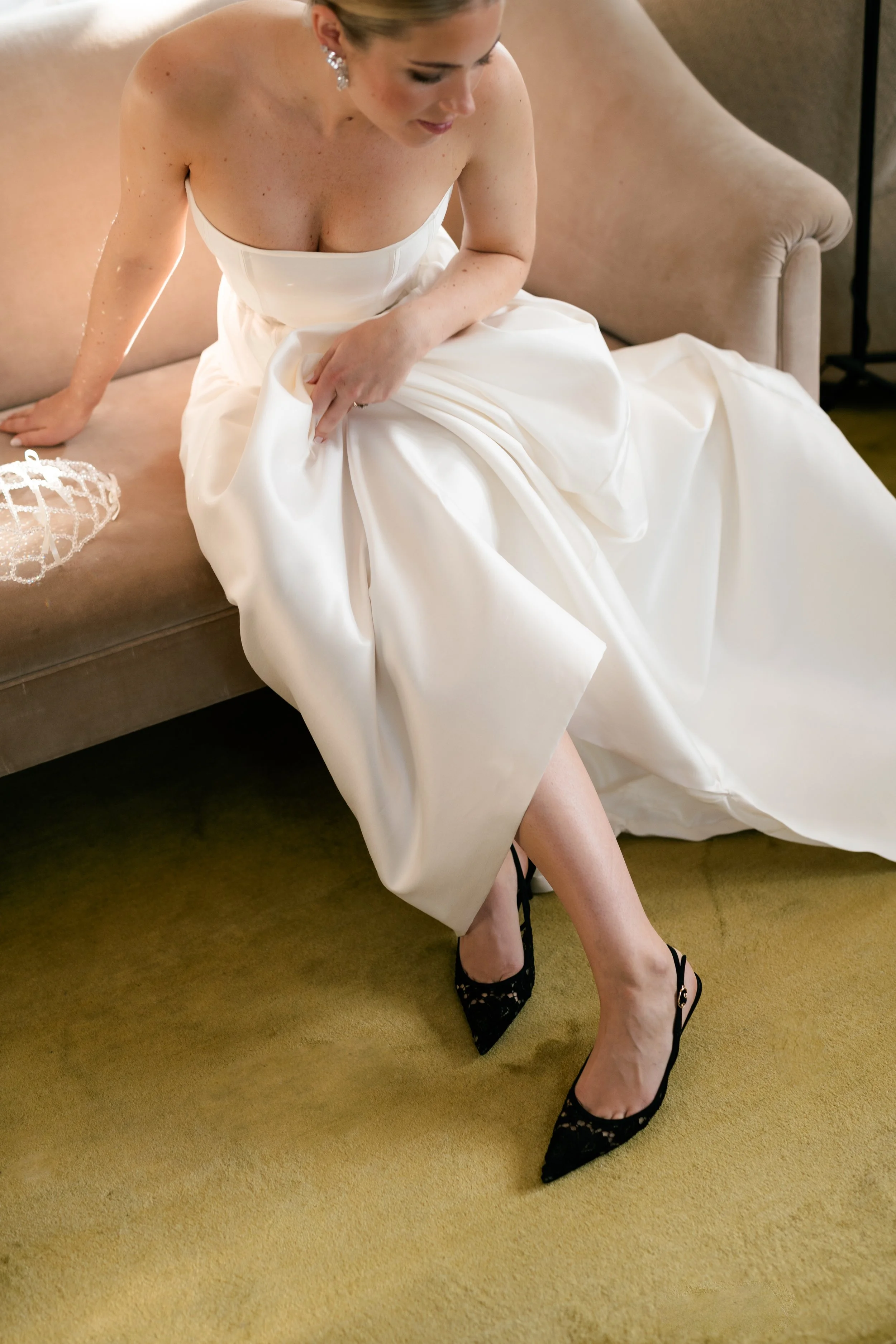 A bride sitting on a beige couch, wearing a white wedding gown and black lace high heels, adjusting her dress.