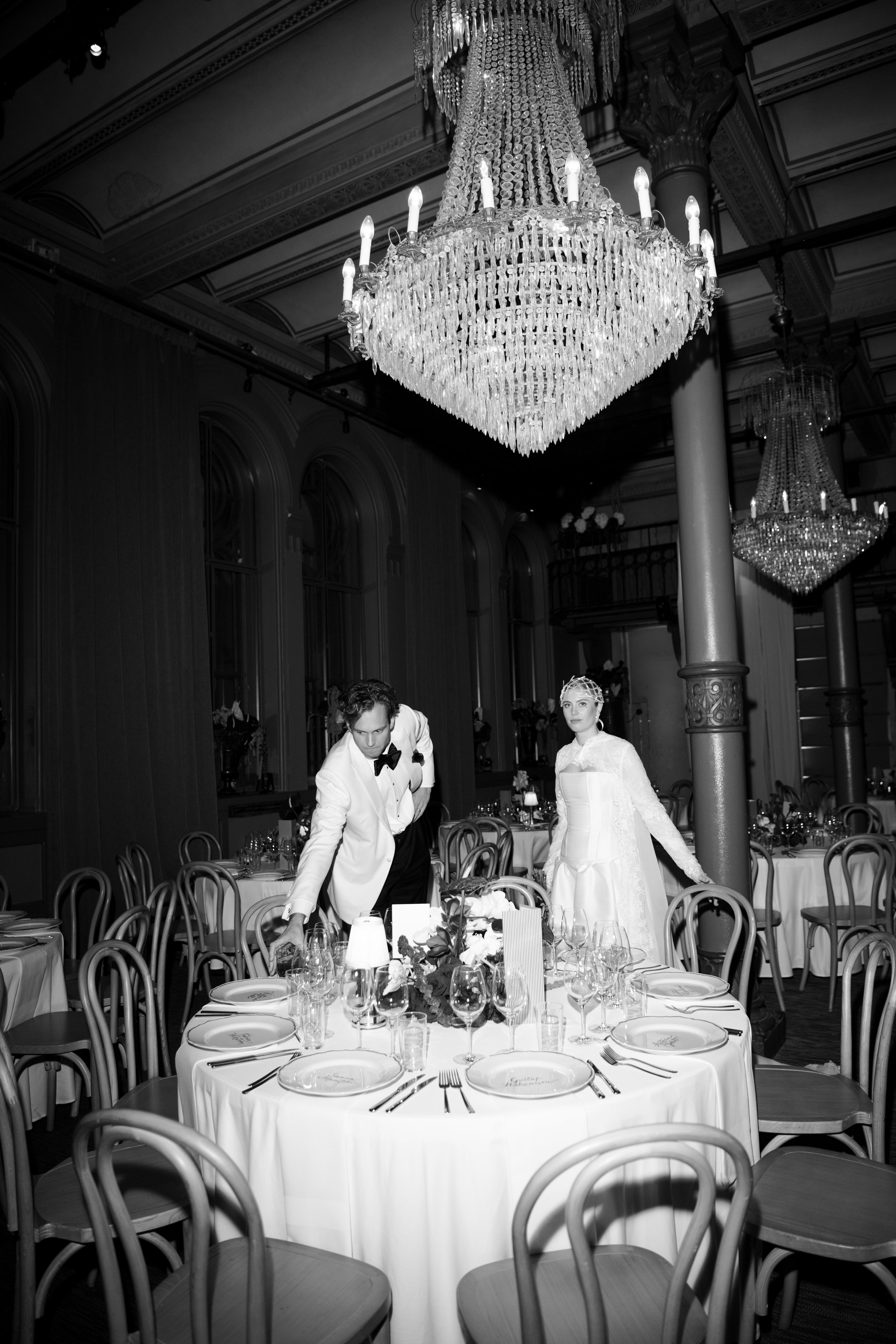 Black and white photo of a luxurious banquet hall with a large chandelier hanging from the ceiling, a table set for a formal event with elegant tableware and floral centerpiece, and a bride and groom in wedding attire standing near the table.