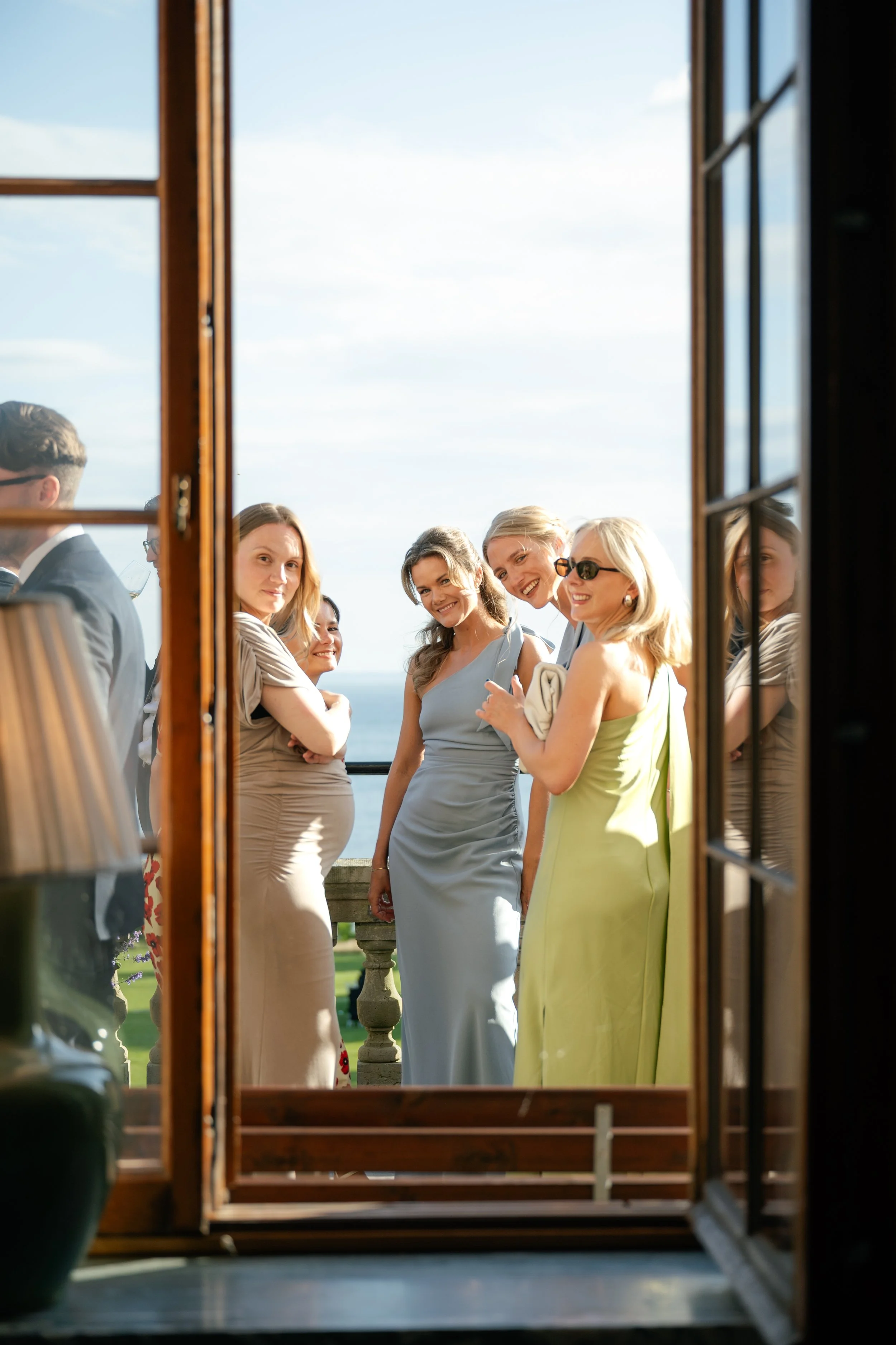 Group of women standing outdoors on a sunny day, viewed through an open window, with a scenic ocean view in the background.