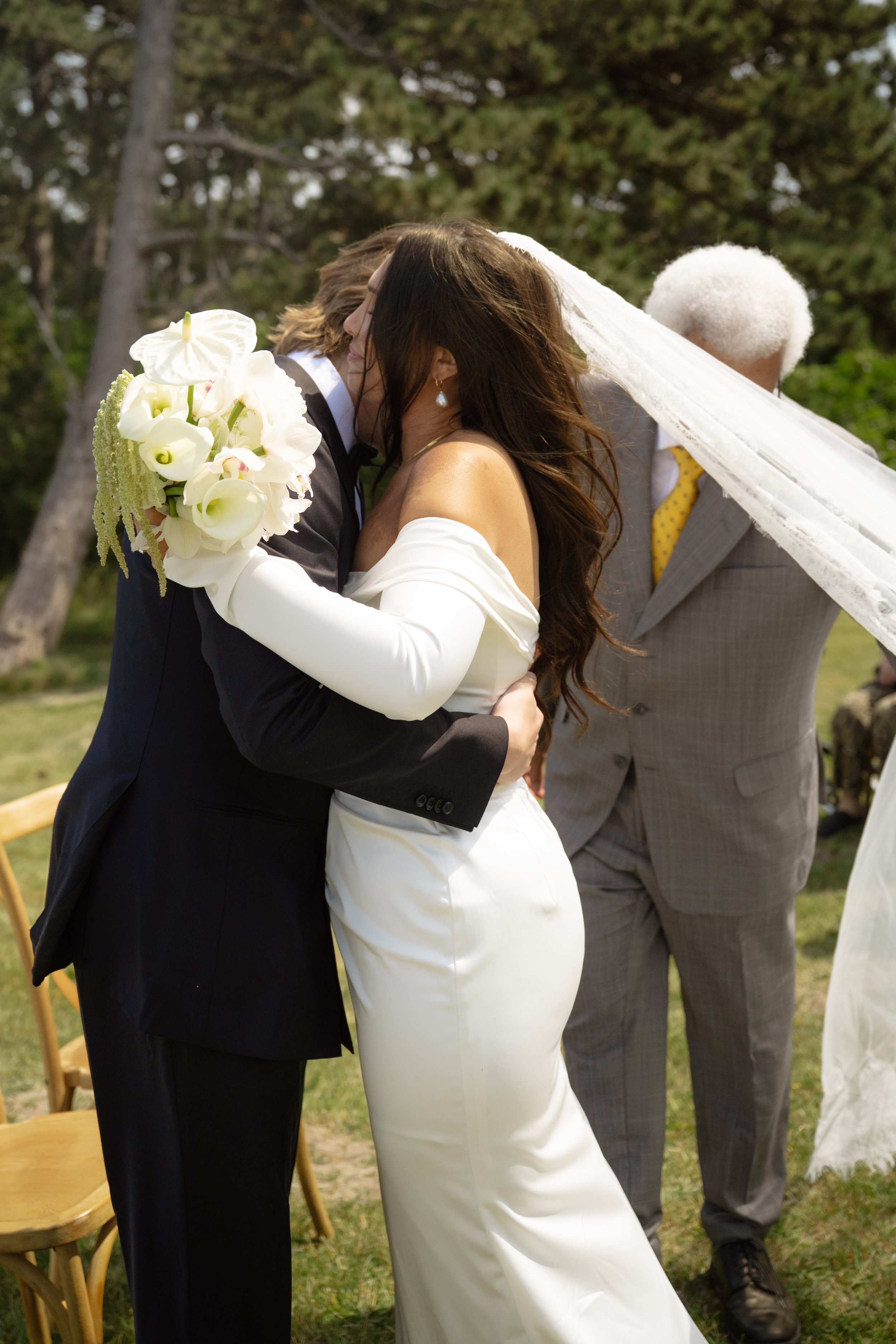Couple kissing at outdoor wedding ceremony, woman in white wedding dress, man in black tuxedo, holding bouquet, officiant in grey suit with yellow tie in background.