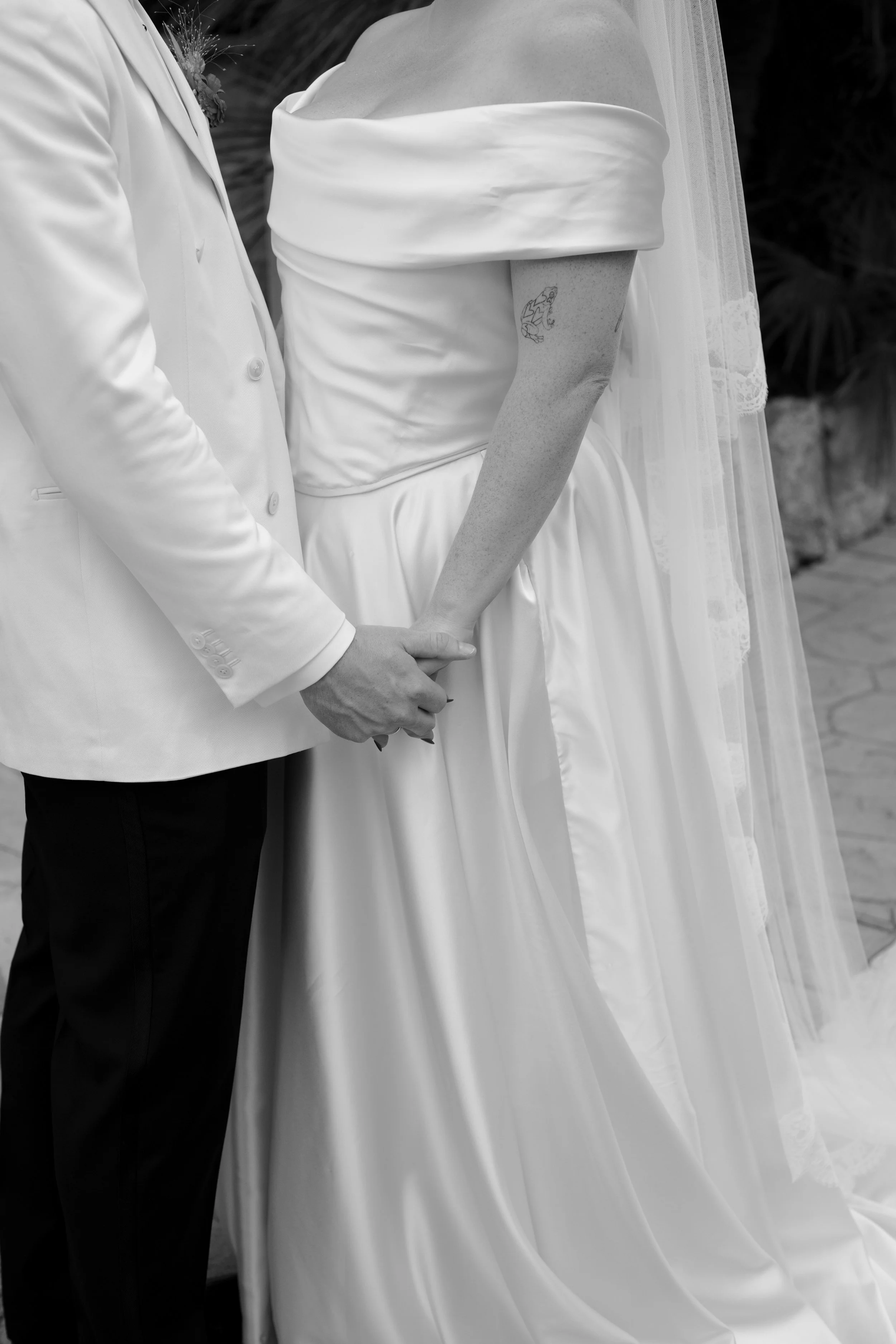 Bride and groom holding hands, with the groom in a white suit and the bride in a white off-shoulder wedding dress, standing outdoors.