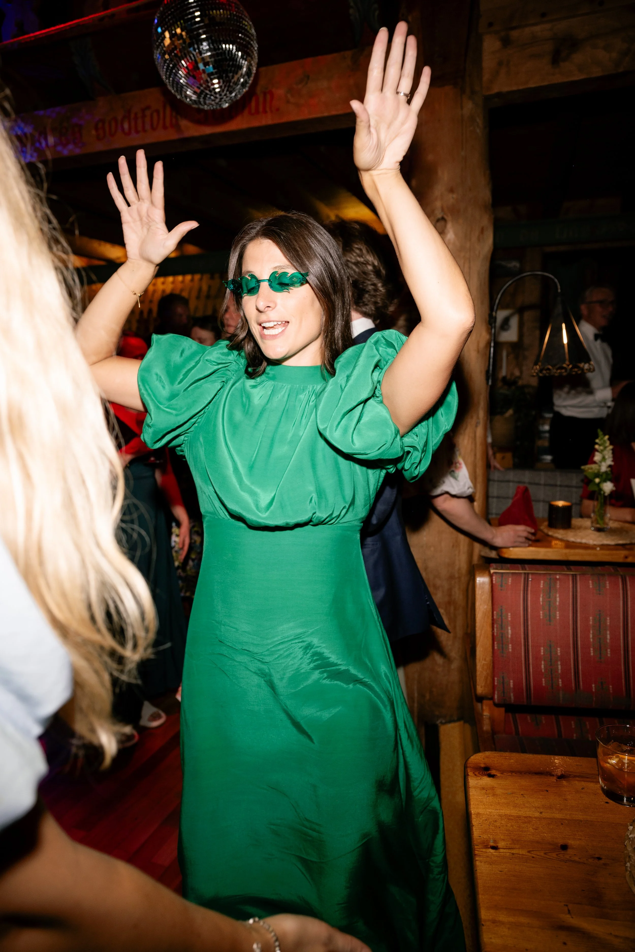 Woman dancing at a party, wearing a bright green dress and sunglasses, with a disco ball overhead.