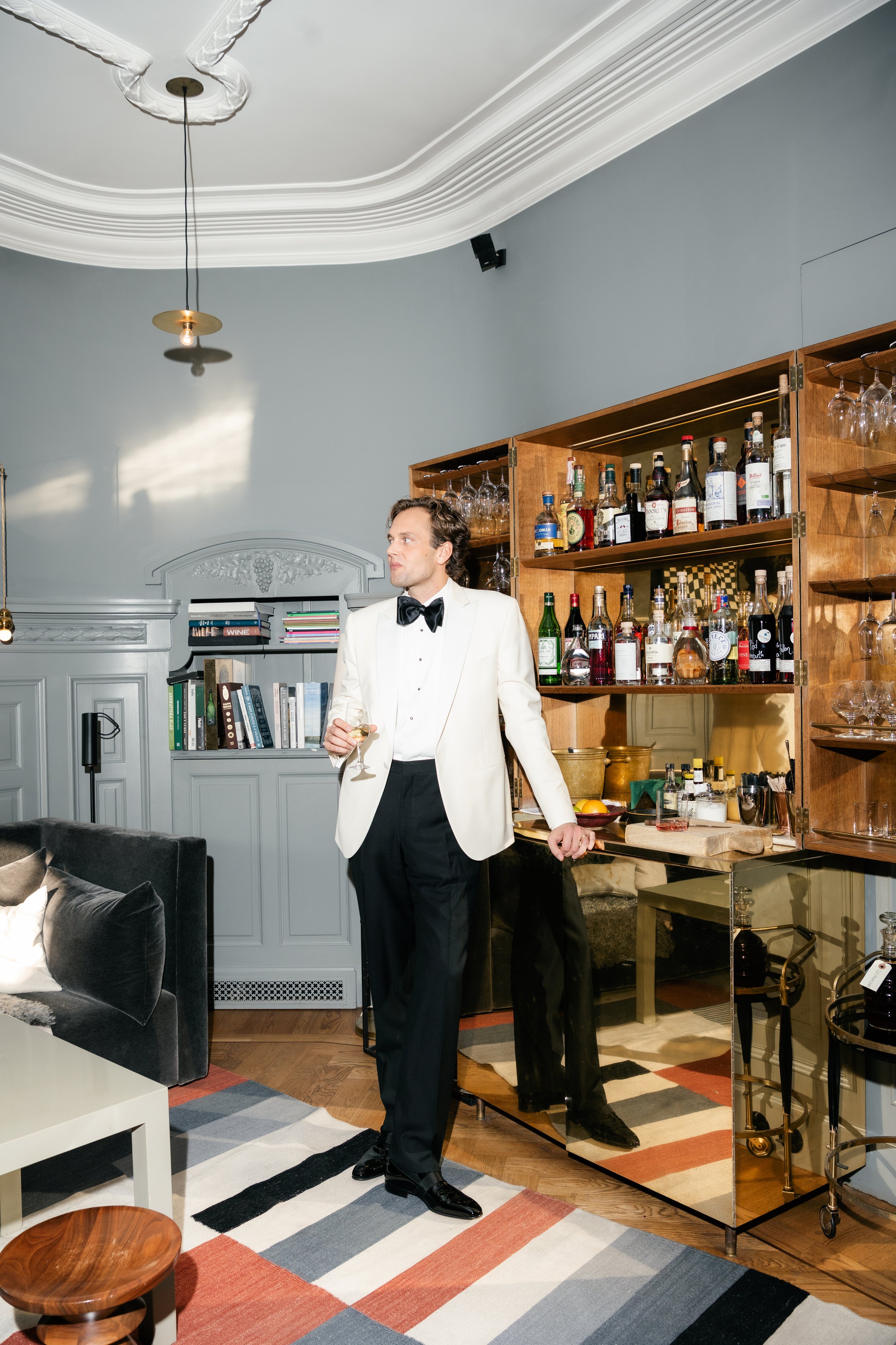 A man in a tuxedo with a white jacket and black bowtie standing next to a bar, holding a glass of wine, in a stylish living room with grey walls, wooden shelving, books, and a colorfully striped rug.