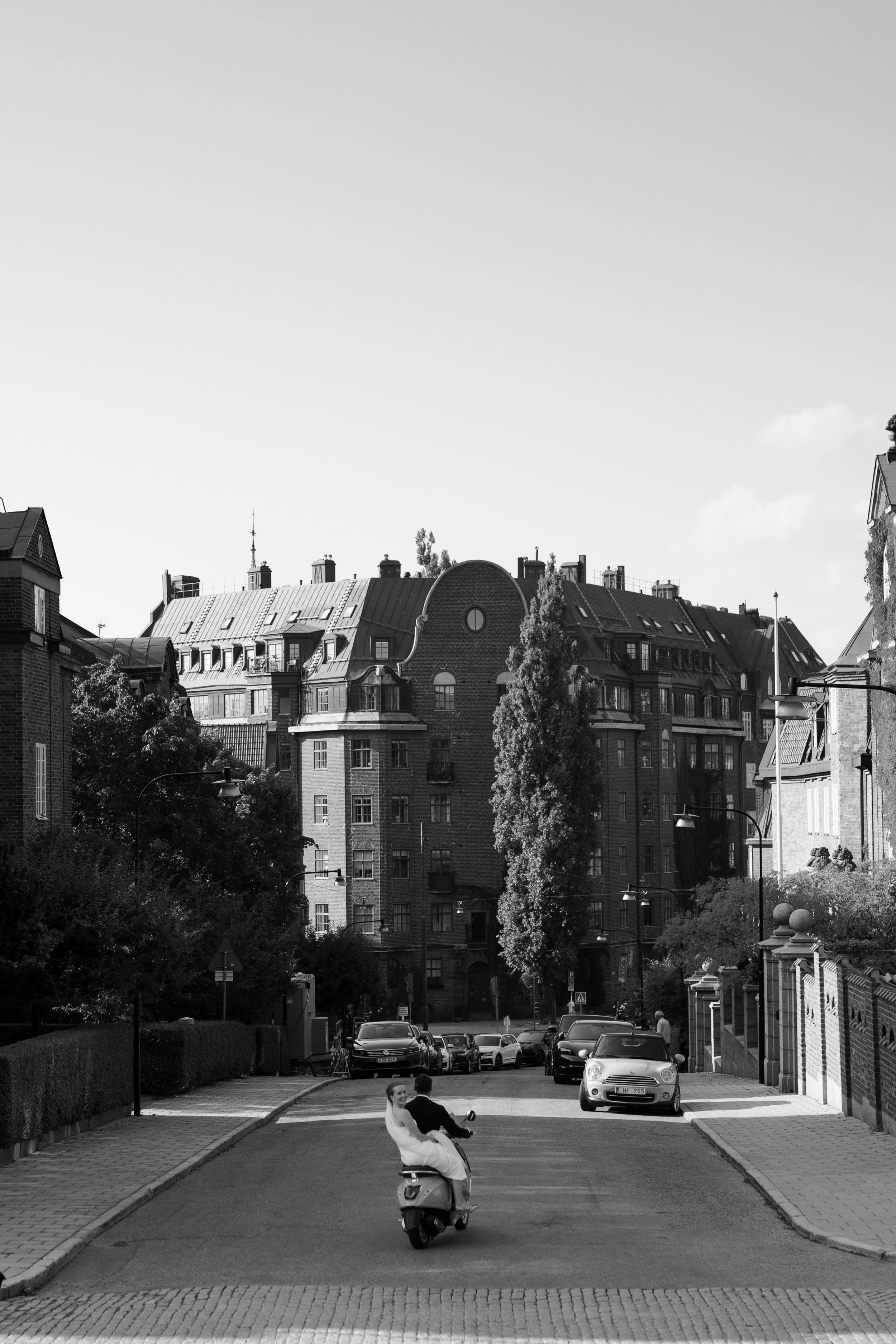 A city street with tall, ornate buildings and trees lining the sidewalk. A couple, dressed in wedding attire, is riding a scooter down the street, with parked cars visible on both sides.