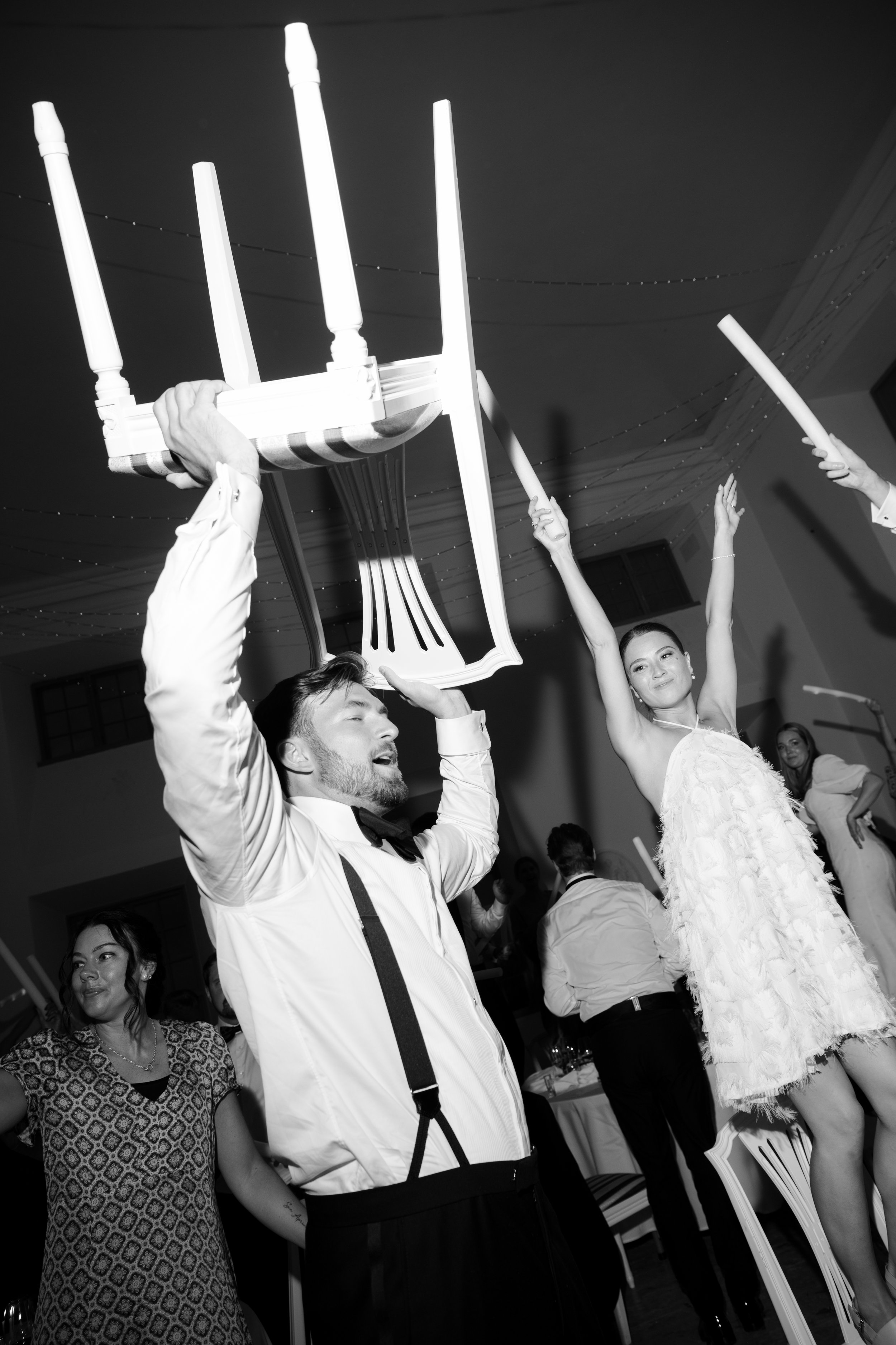 Group of people at a celebration, some holding chairs above their heads, facing upward, with a woman in a feathered dress and a man in suspenders and a bow tie in the foreground.