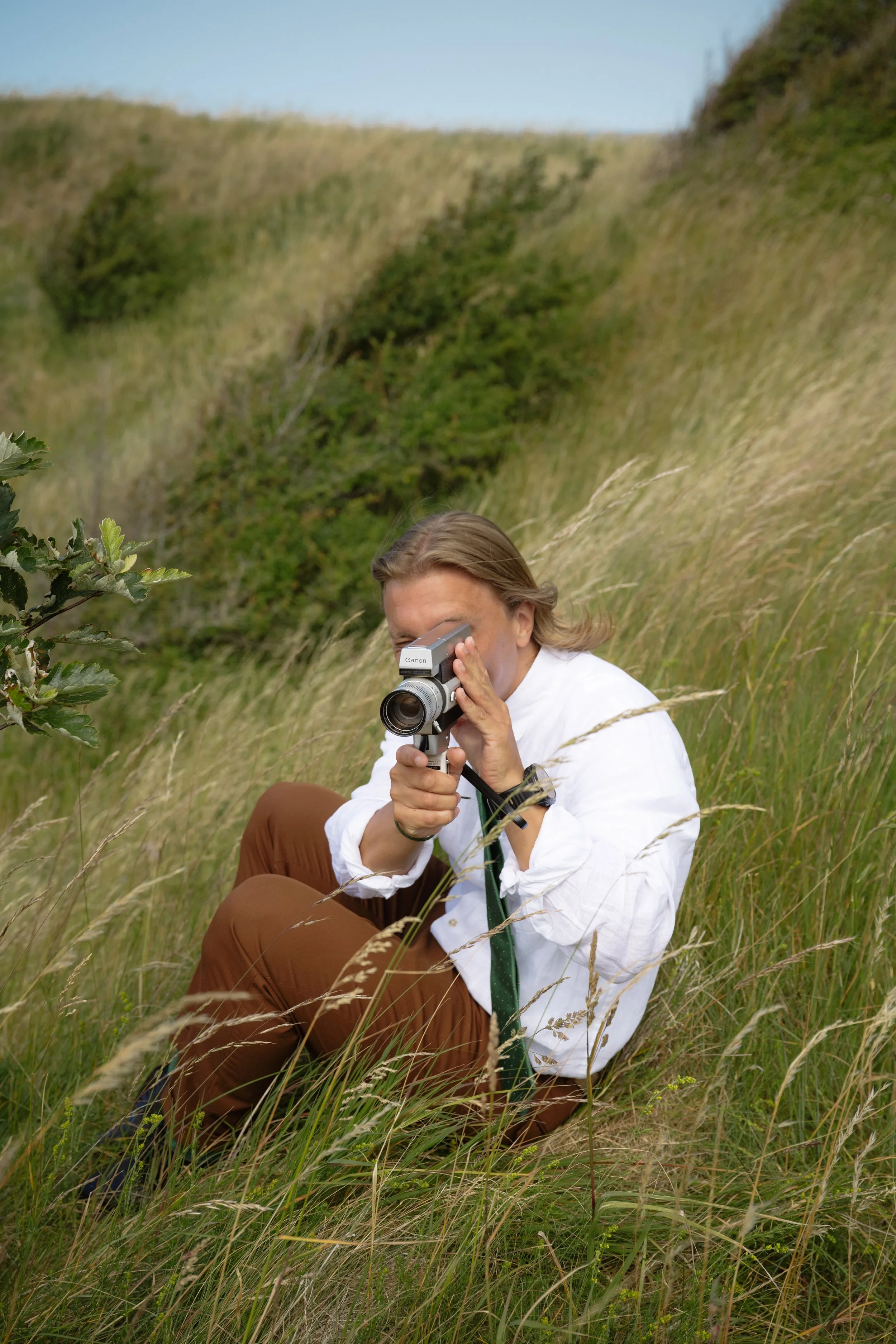 A woman sitting in tall grass on a hillside, taking a photograph with a vintage camera.