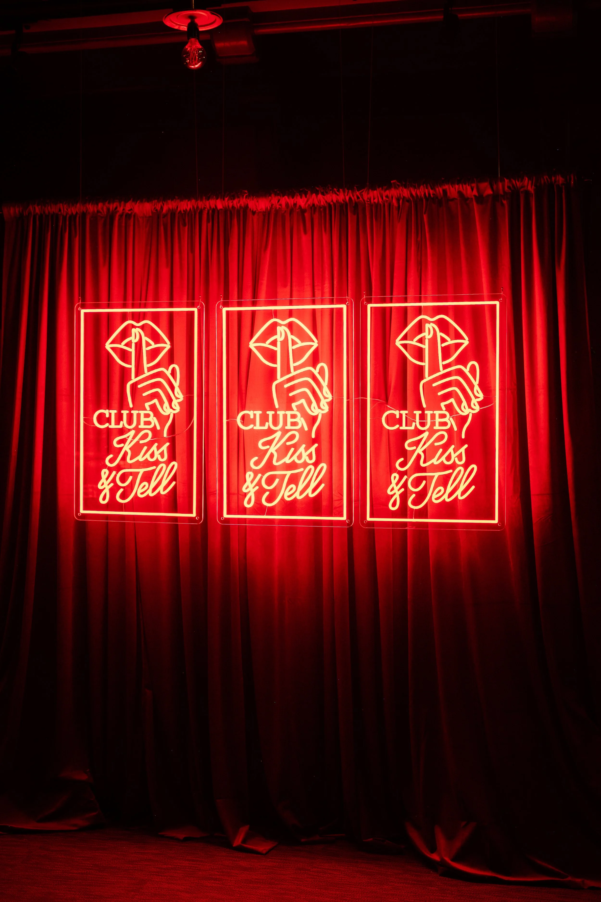 Neon signs that say 'Club Kiss & Tell' with a graphic of lips and a finger over them, hanging on red curtains.