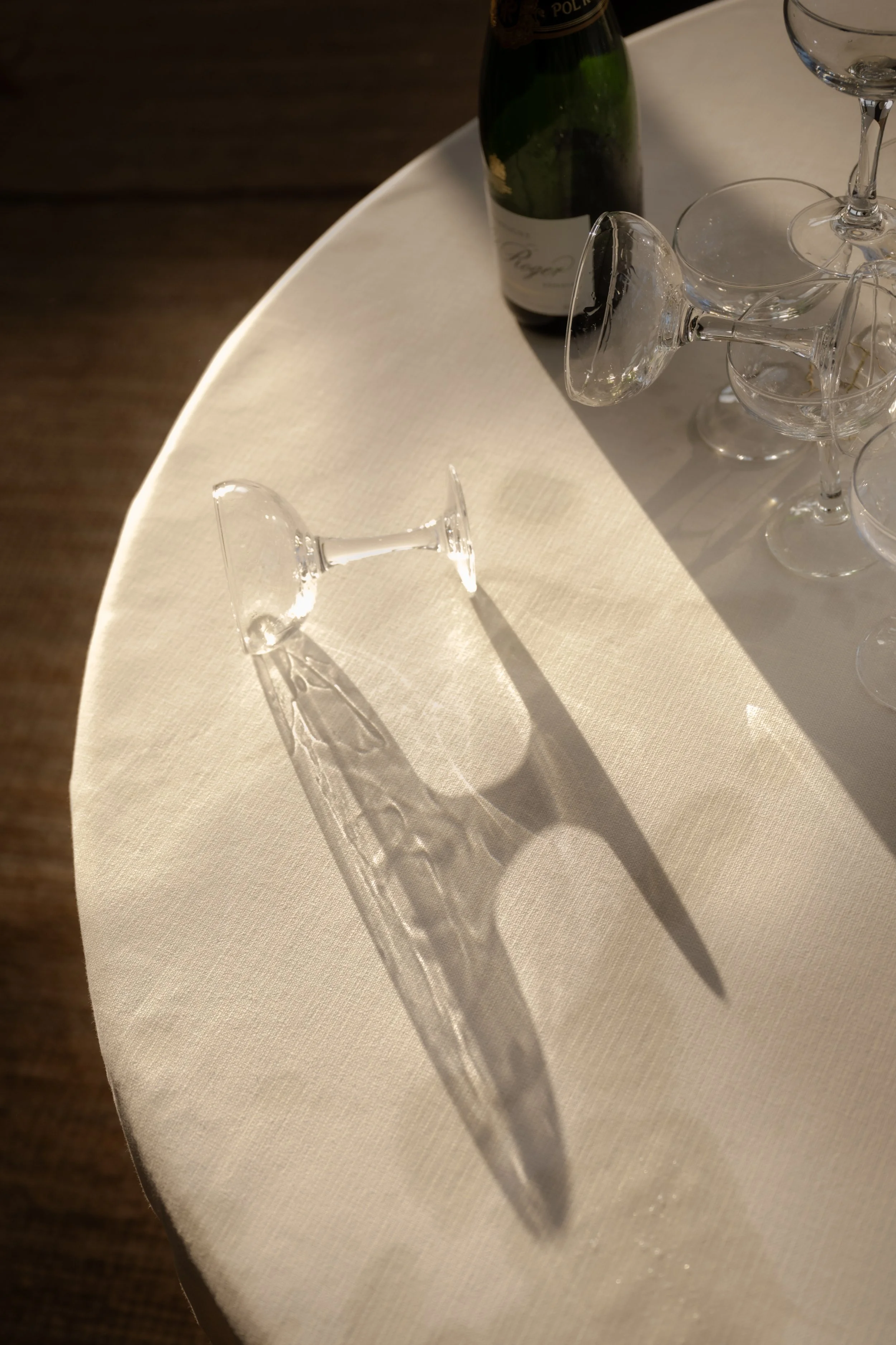 A white tablecloth-covered table with an upside-down wine glass casting a shadow in sunlight, next to a partially visible champagne bottle and several other empty glasses.