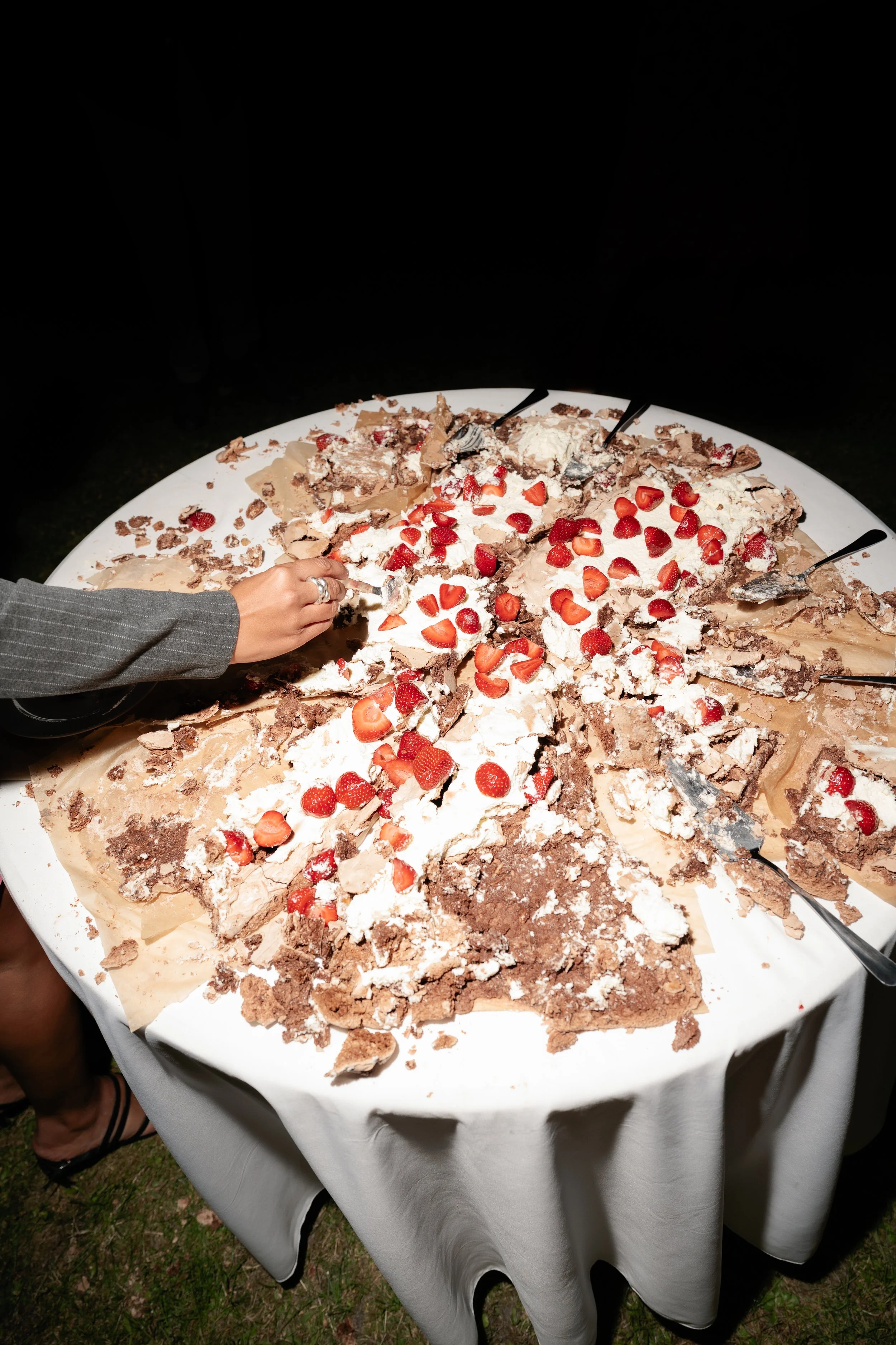 A large, half-eaten chocolate and vanilla cake with strawberries, whipped cream, and chocolate shavings on a white tablecloth, with a person's hand reaching for a piece at a celebration or party.
