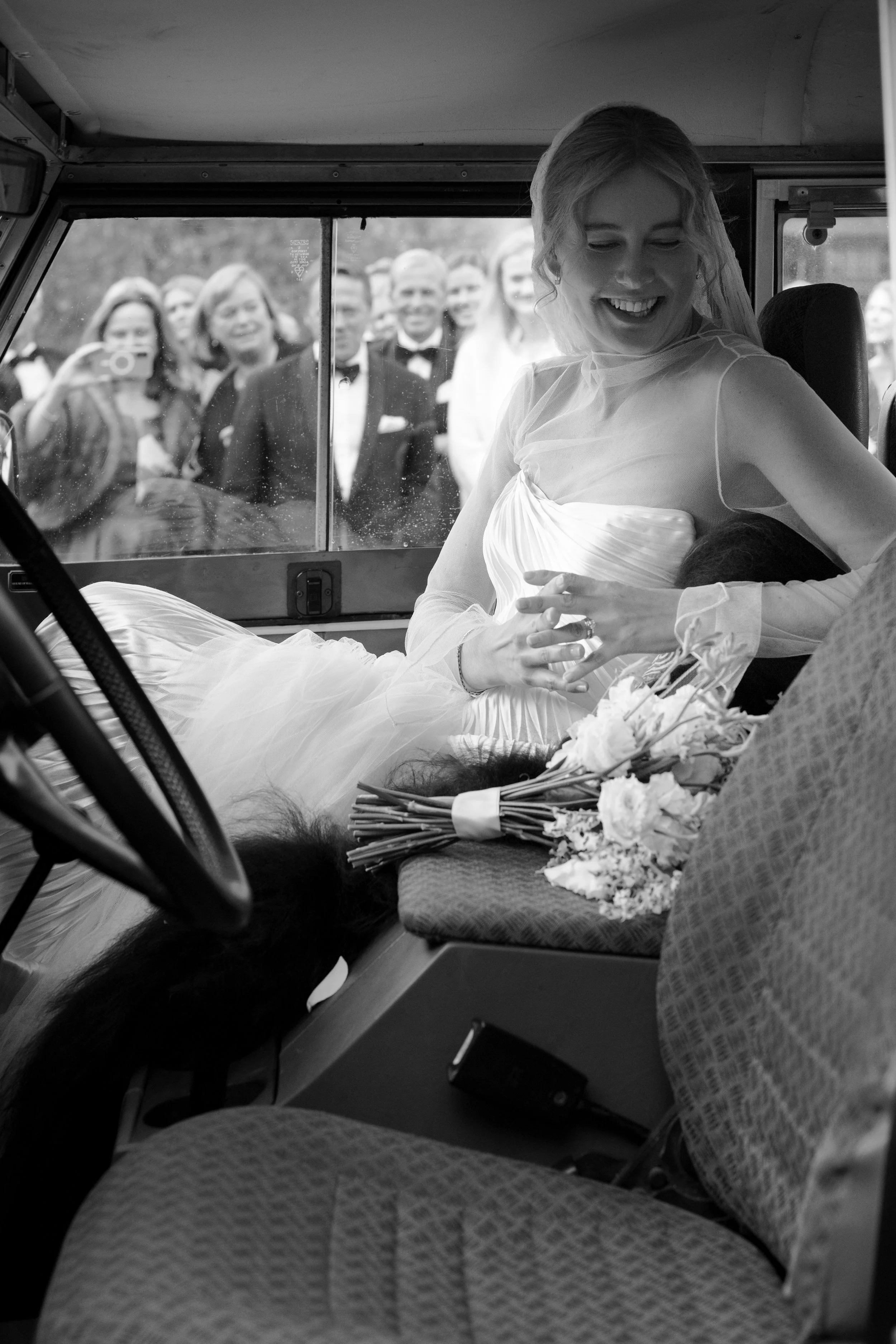 A bride in a wedding dress sitting in a vehicle, smiling and holding flowers, with wedding guests outside taking photos through the window.