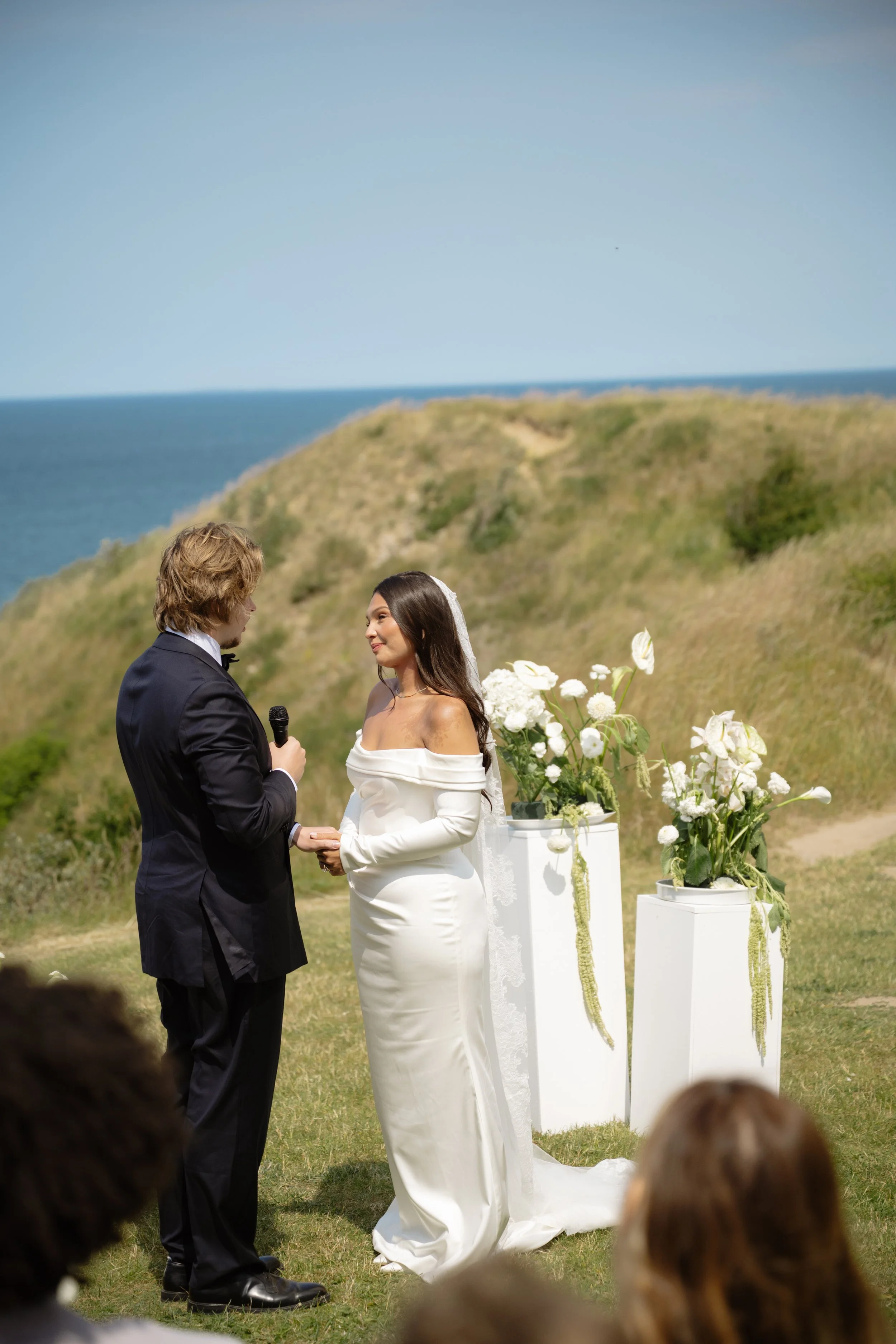 A bride and groom exchanging vows during an outdoor wedding ceremony by the ocean on a sunny day.