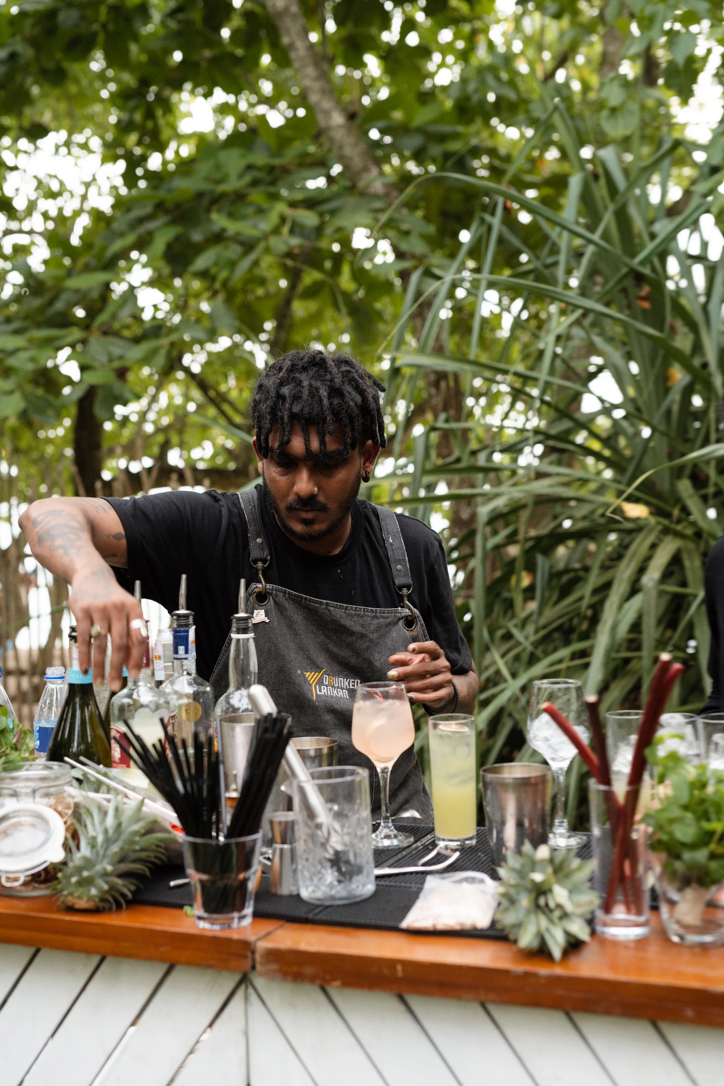 A bartender mixing drinks outdoors, surrounded by tropical plants and a variety of cocktail ingredients and garnishes on the bar.