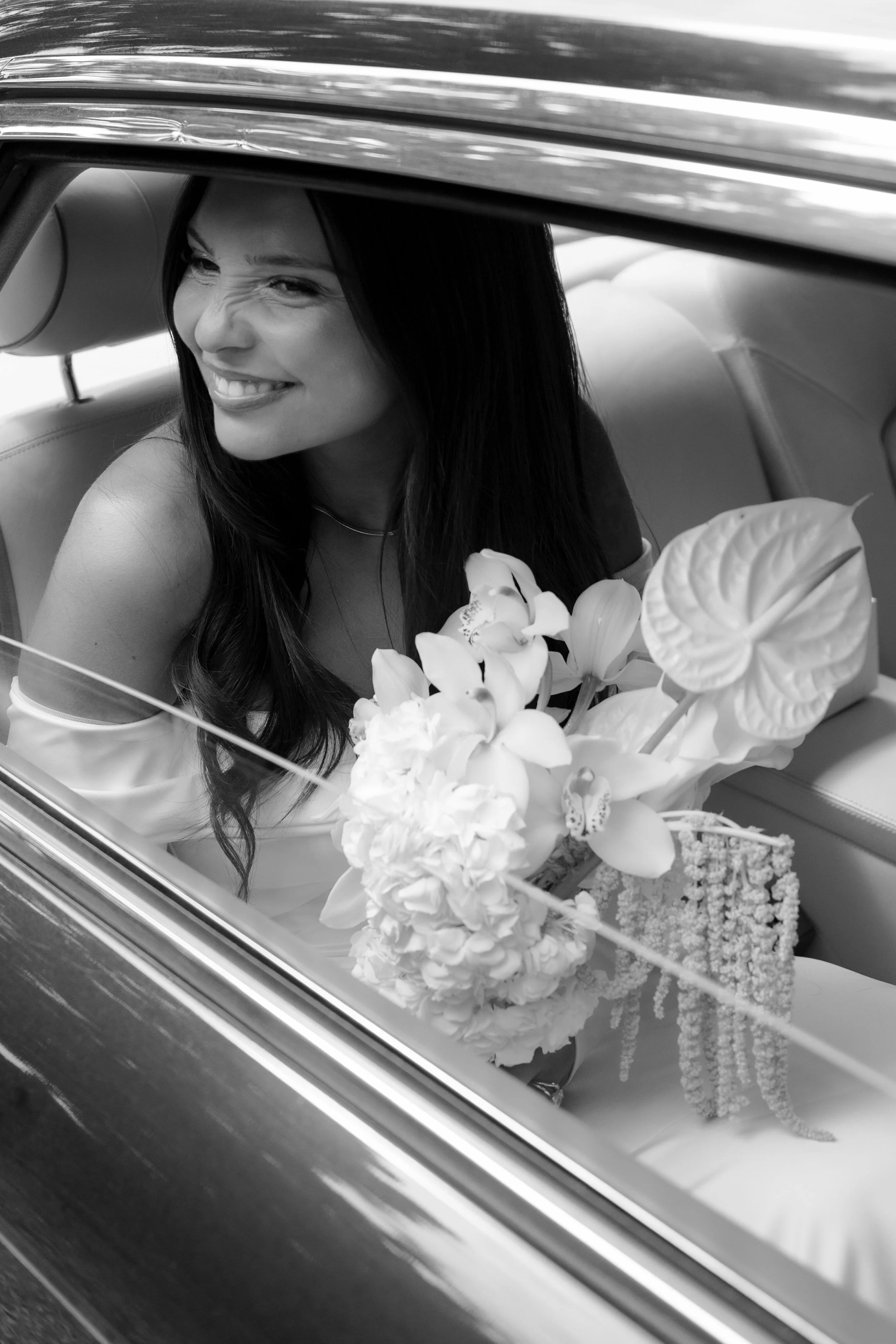 A woman sitting in a vintage car, smiling and winking while holding a bouquet of flowers.