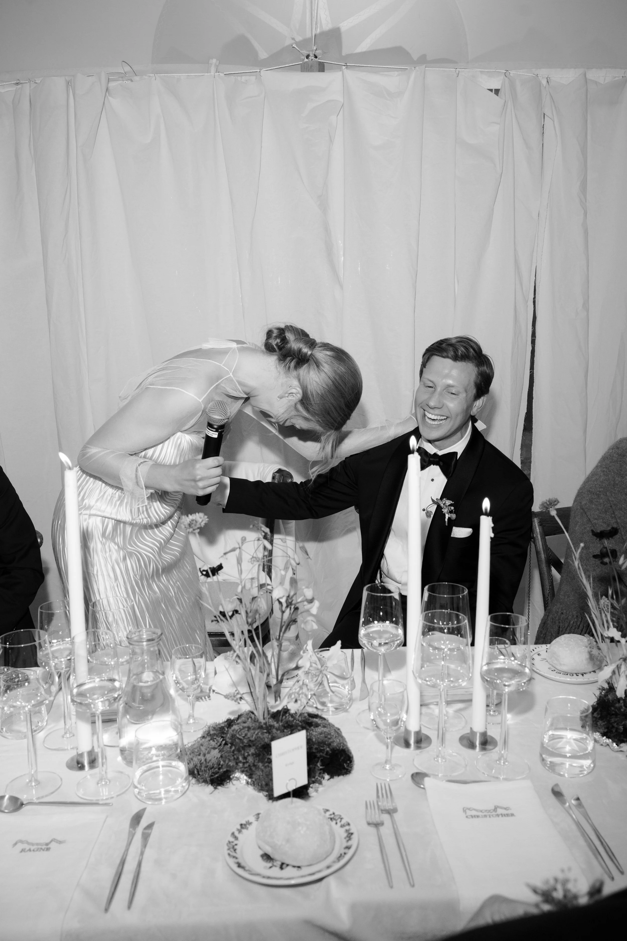 A black and white photo of a wedding reception with a woman in a dress holding a microphone leaning towards a smiling man in a tuxedo at the table, with candles, glasses, and floral arrangements on the table.