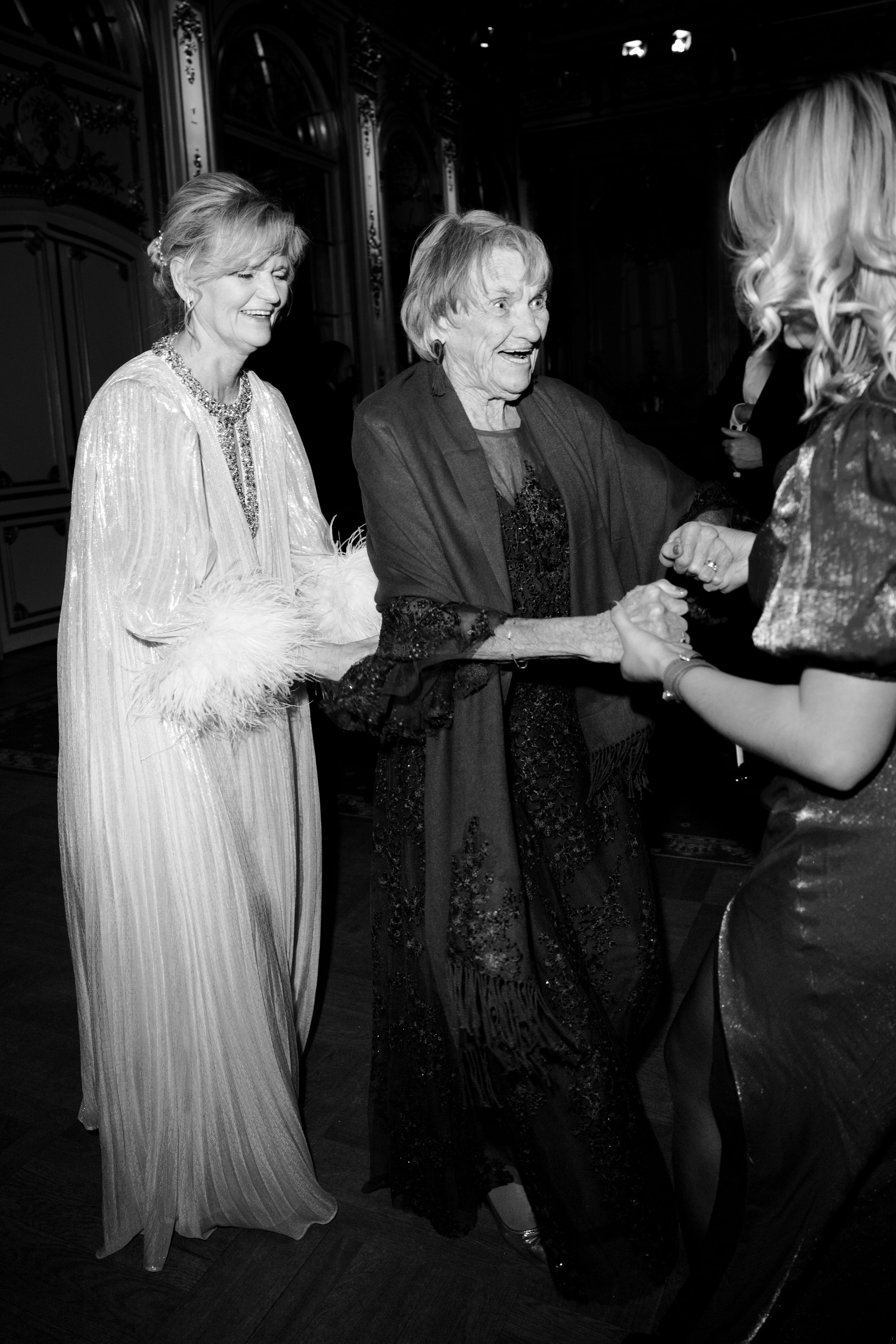 Three women at a formal event holding hands, smiling and dancing in an elegant room.