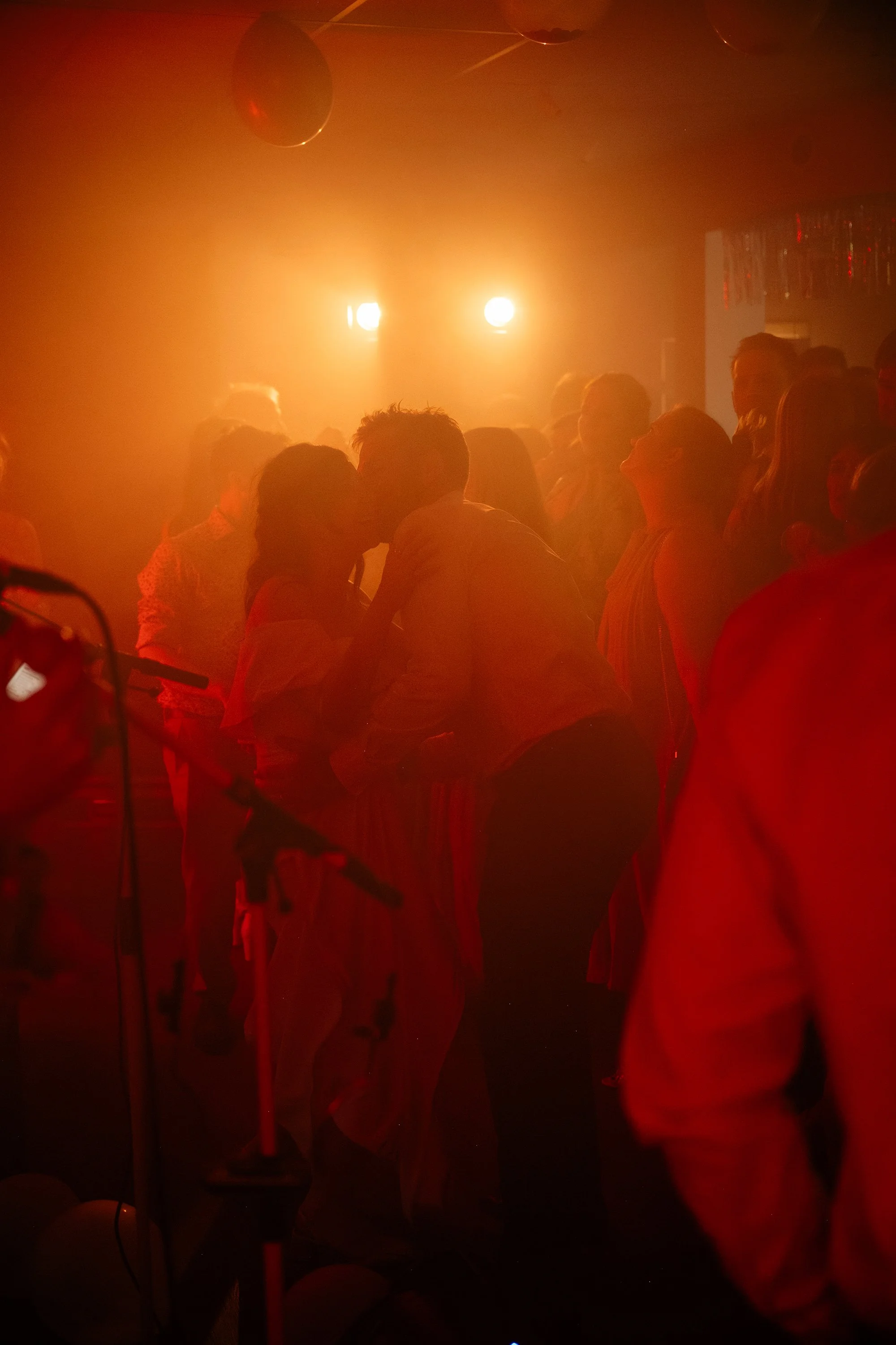 Couple sharing a kiss on a dance floor illuminated with red and orange lights, surrounded by other people dancing in the background.