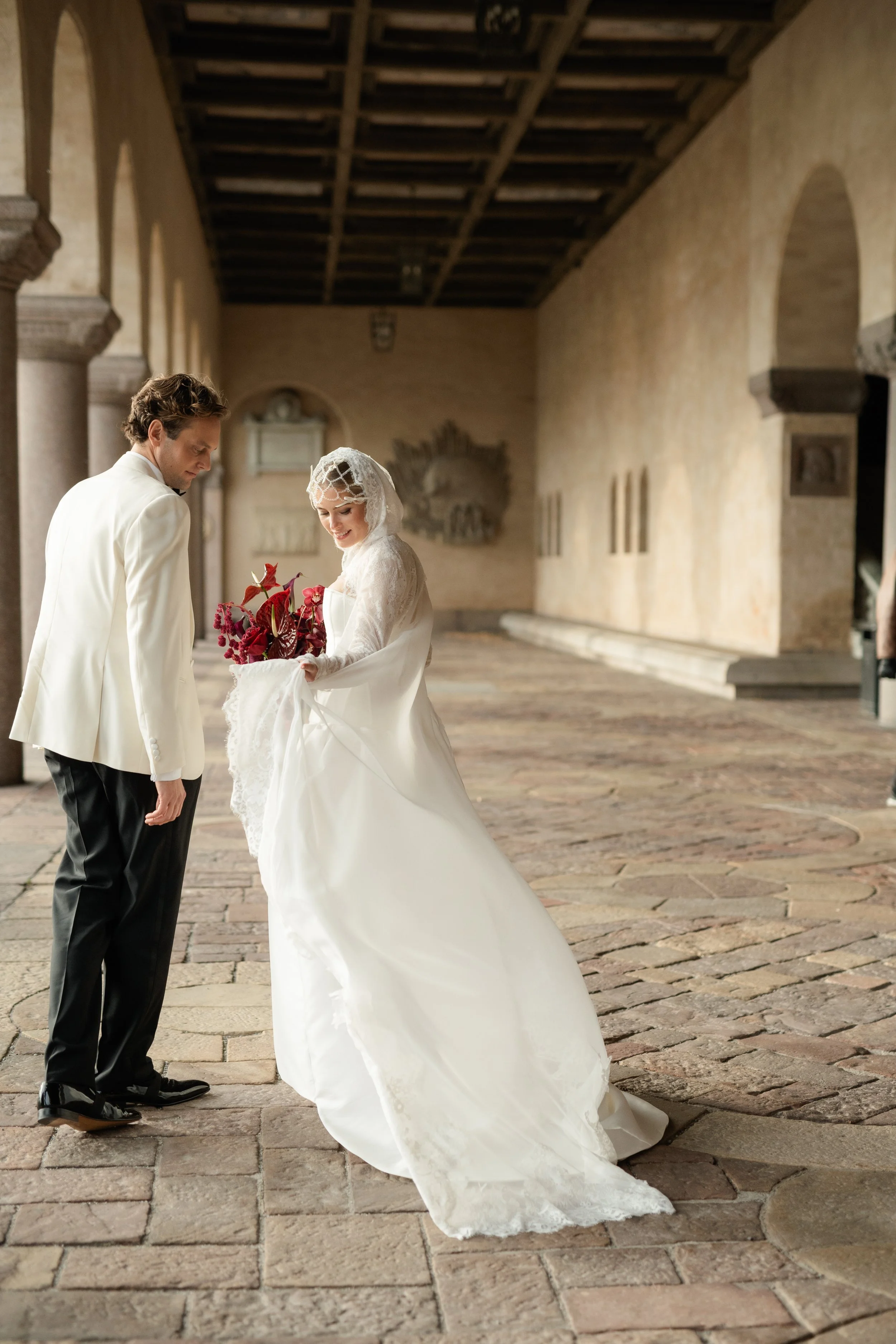 A bride in a white wedding dress holding a bouquet of red flowers, standing in an old stone corridor with a groom in a white tuxedo looking down at her.