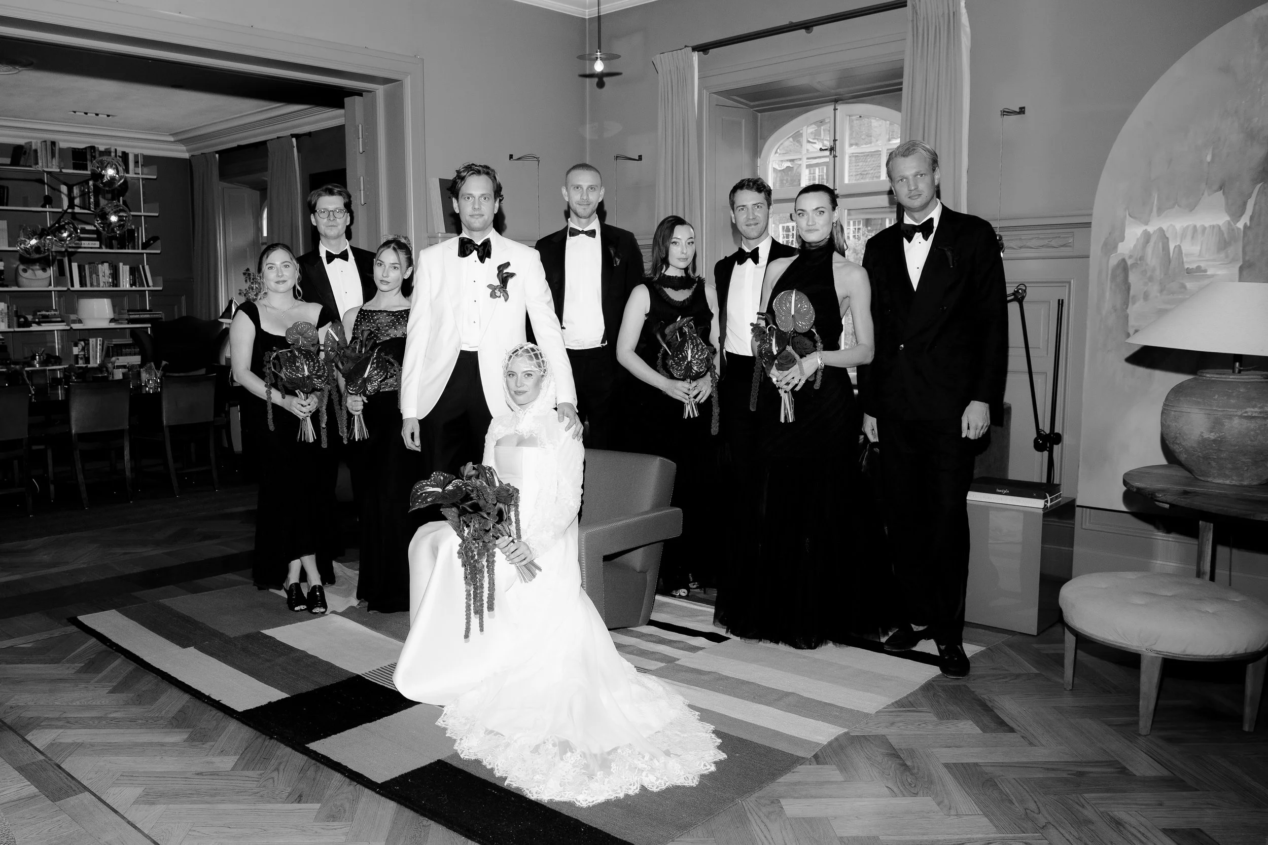 A group of eleven people dressed in formal attire posing for a photo indoors, with one woman in a wedding dress sitting on a chair in the front. The others are standing behind her, some holding bouquets, in what appears to be a wedding celebration or