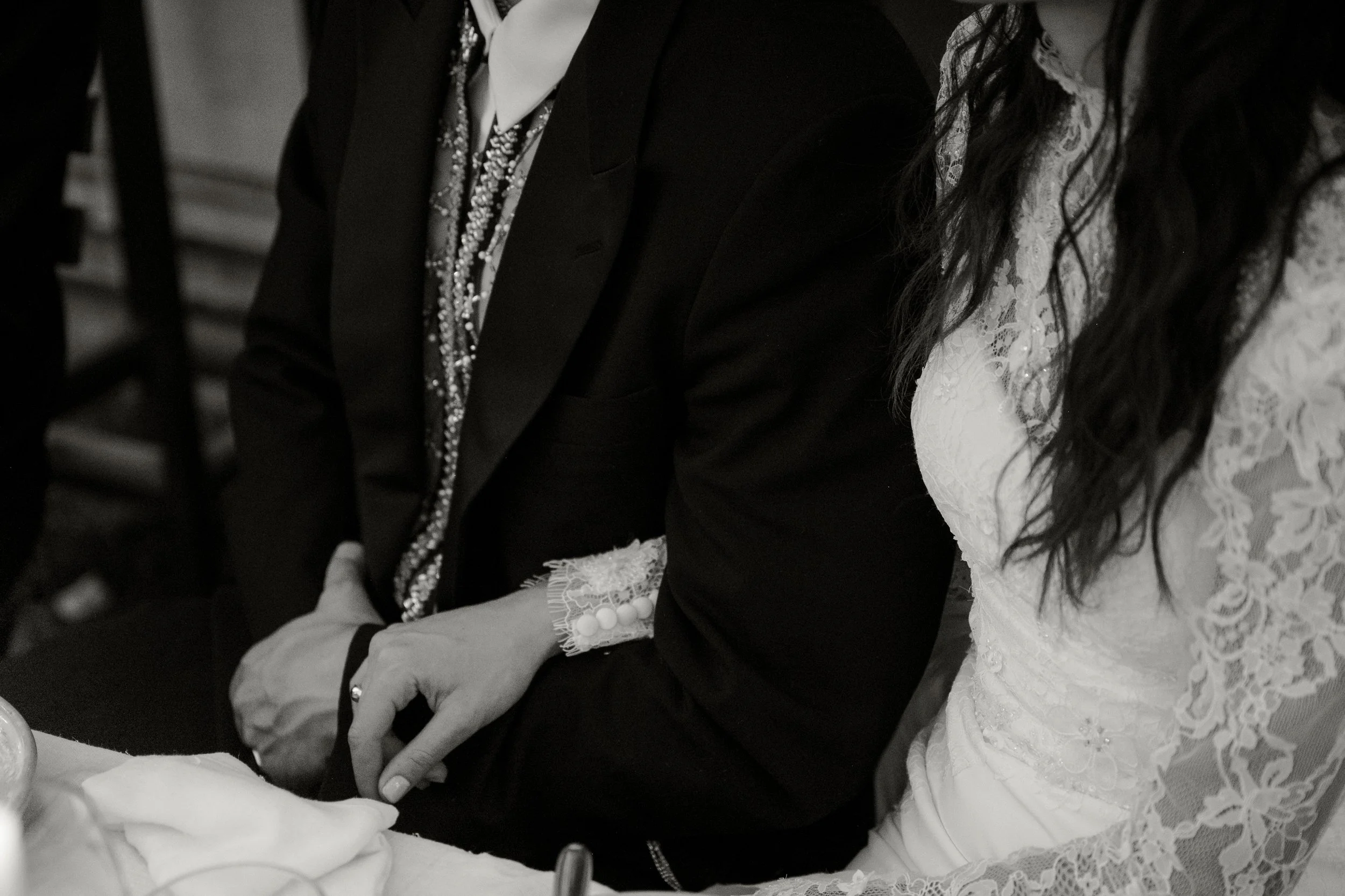 Close-up of a bride and groom sitting next to each other at a wedding reception, with the bride wearing a lace dress and the groom in a dark suit.