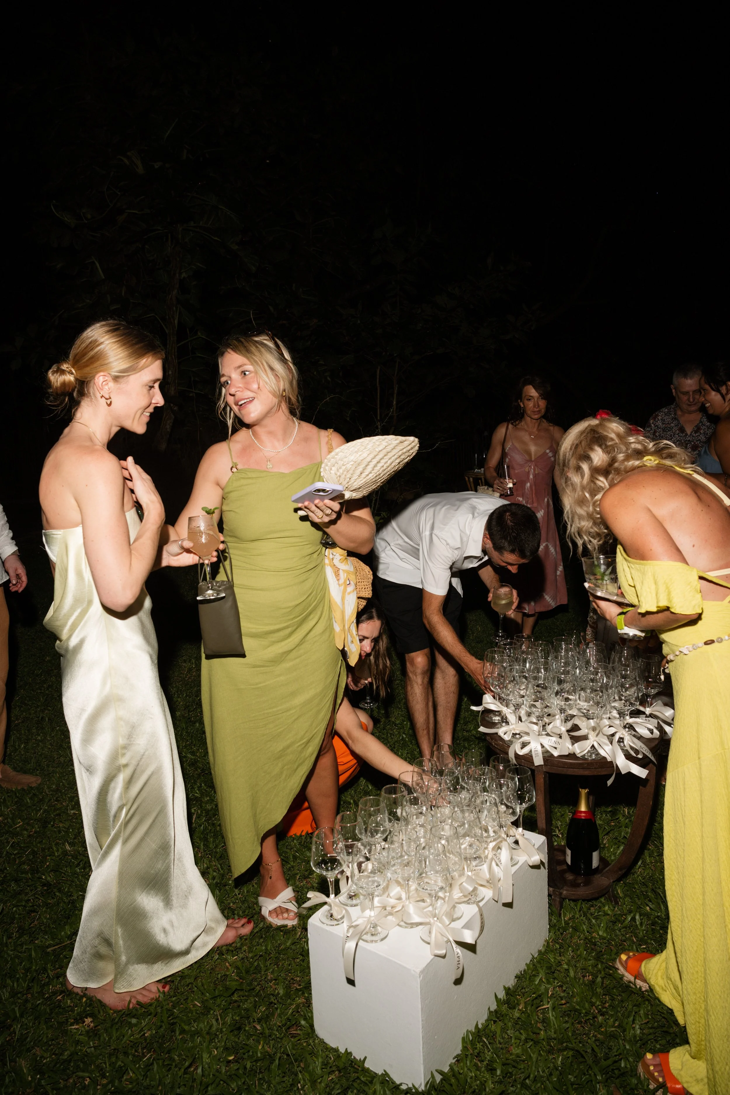 People at a nighttime outdoor celebration exchanging drinks and selecting glassware from a table decorated with ribbons.