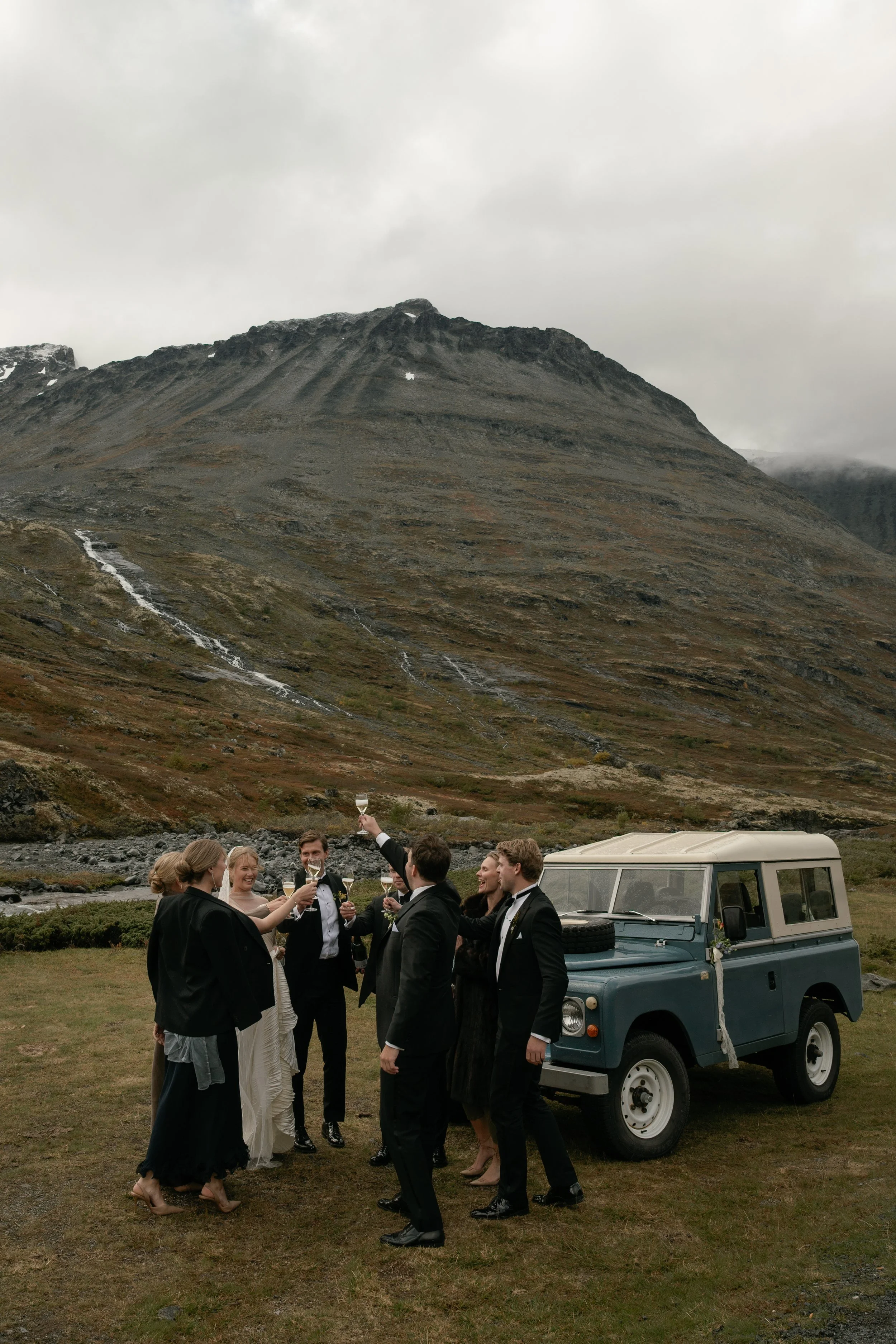 A group of people in formal attire celebrating with drinks outdoors near a vintage vehicle at the foot of a mountain with waterfalls on a cloudy day.