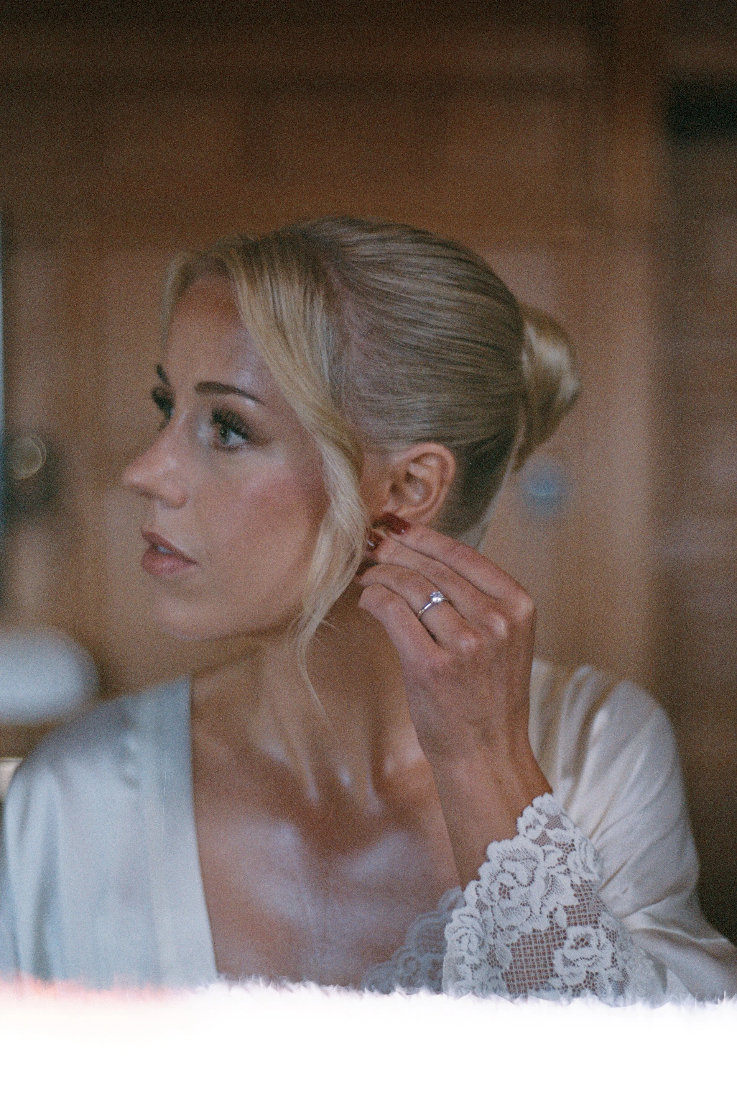 A woman with blonde hair styled in an elegant updo is putting on an earring, wearing a satin robe with lace detail on the sleeves, and a ring on her finger.