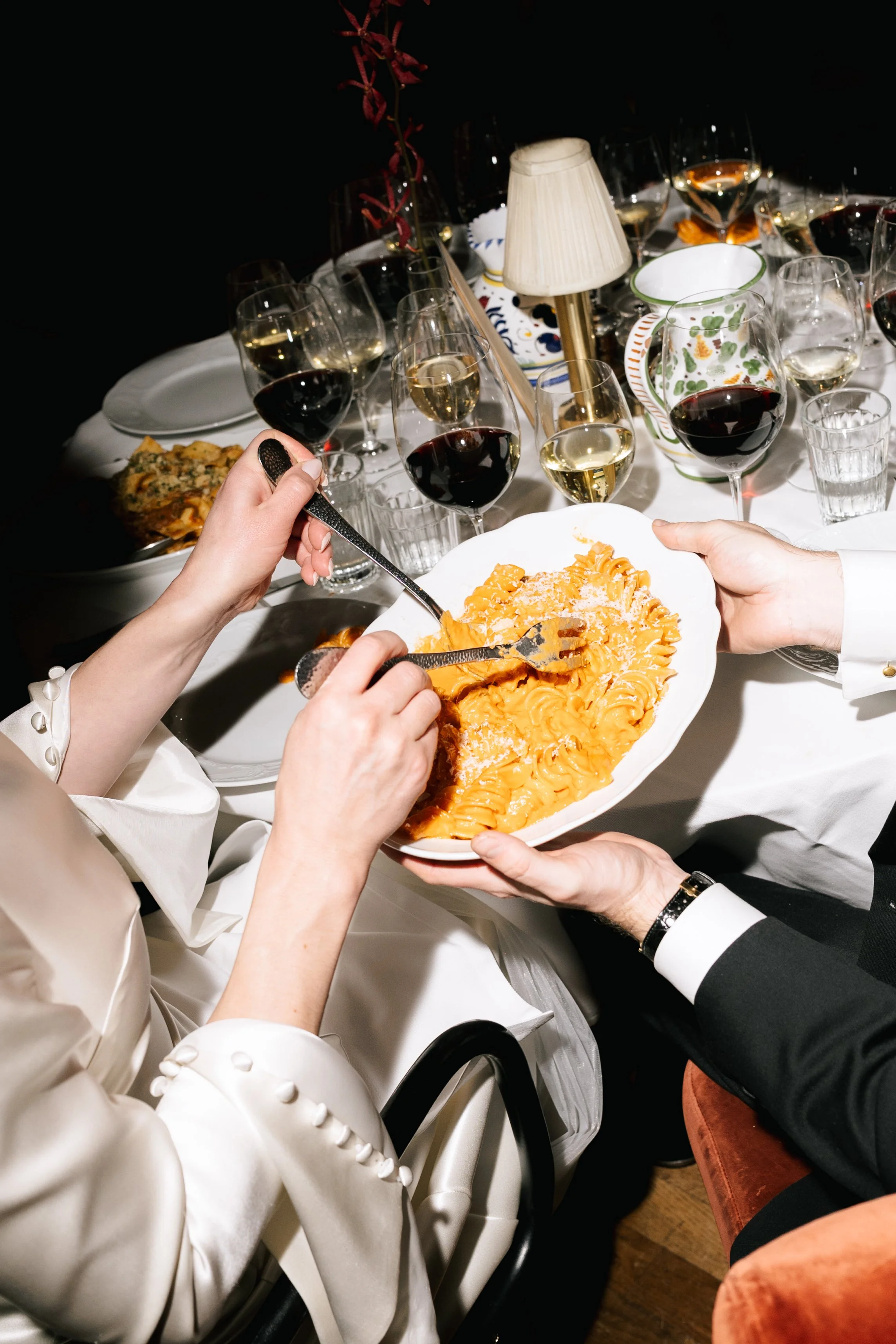 People sharing a plate of spaghetti at a dinner party with wine glasses and a decorated table.