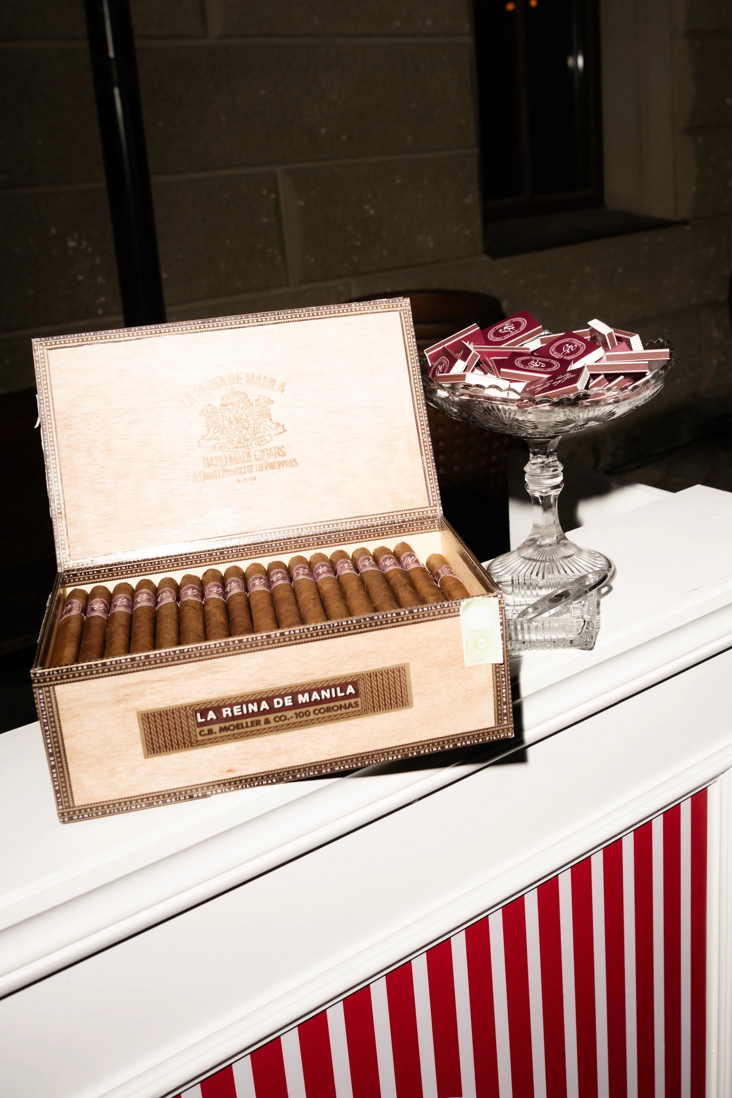 Open box of cigars labeled 'La Reina de Manila' on a white table, next to a glass bowl filled with small rectangular boxes, with a red and white striped surface below.