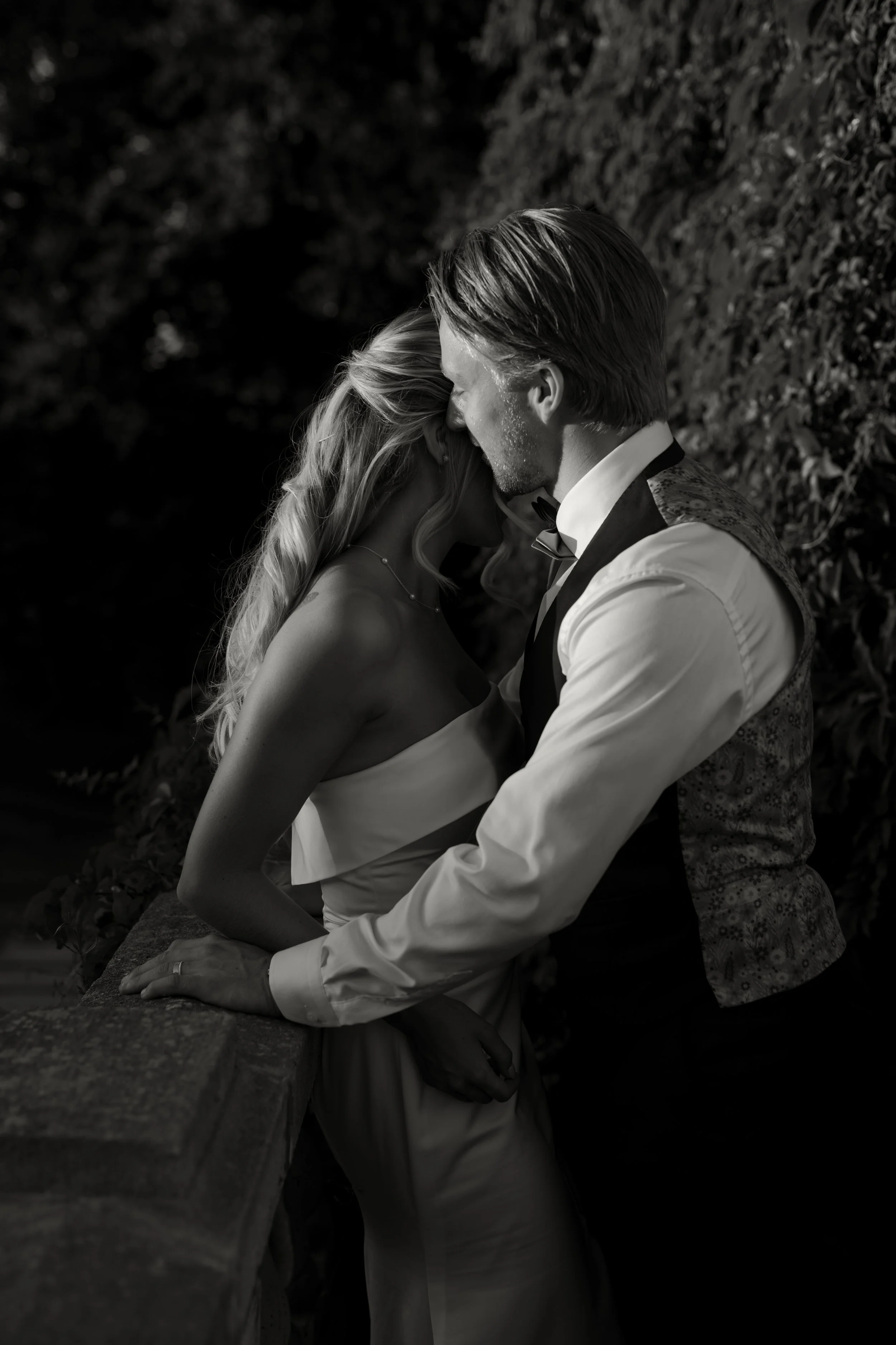 A black and white photo of a couple on their wedding day, with the bride and groom close together, touching foreheads and holding hands, outdoors near a stone railing.