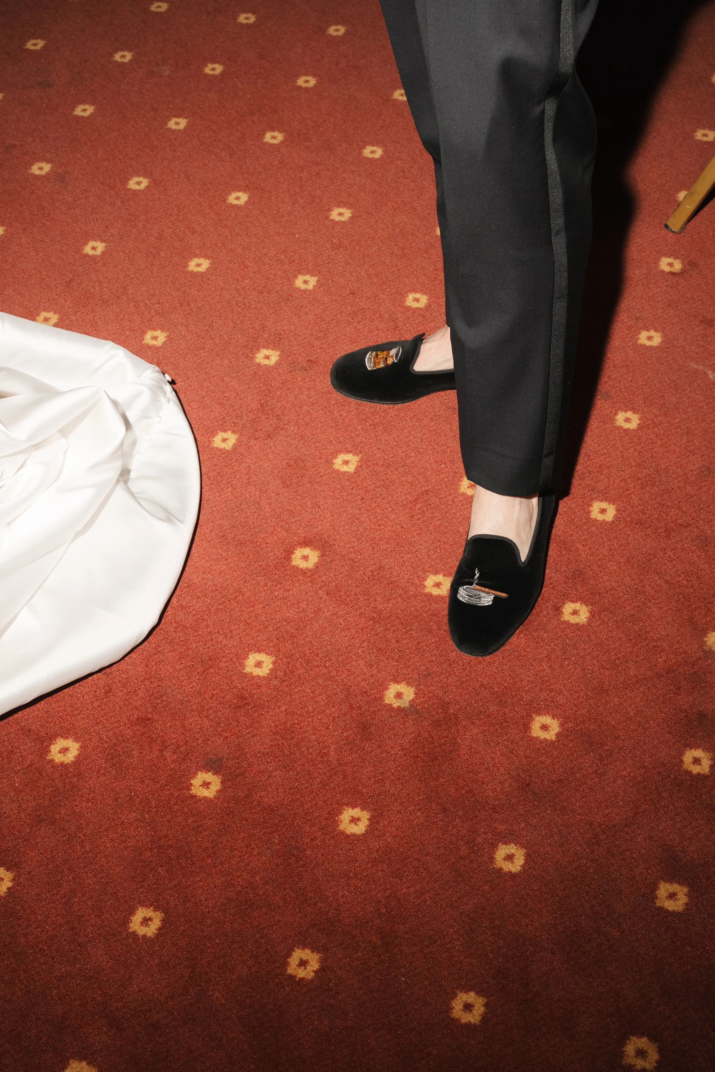 Person standing on red carpet wearing black shoes with embroidered designs, black pants, next to a white cloth-covered object or table.